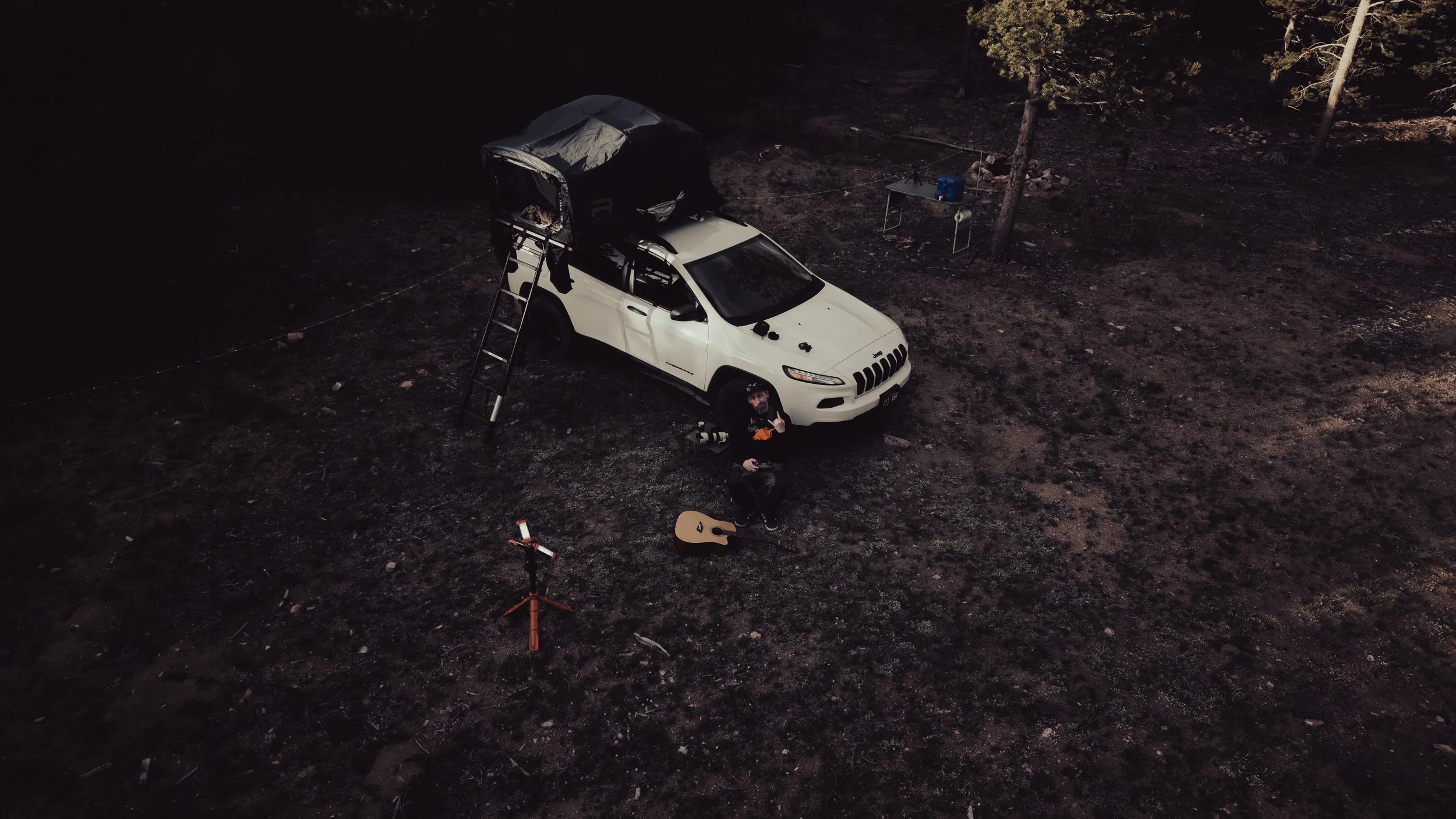 Nighttime outdoor scene with a white SUV parked on dirt, a person sitting with a guitar, a ladder up to a rooftop tent, and a small table with camping supplies surrounded by trees.
