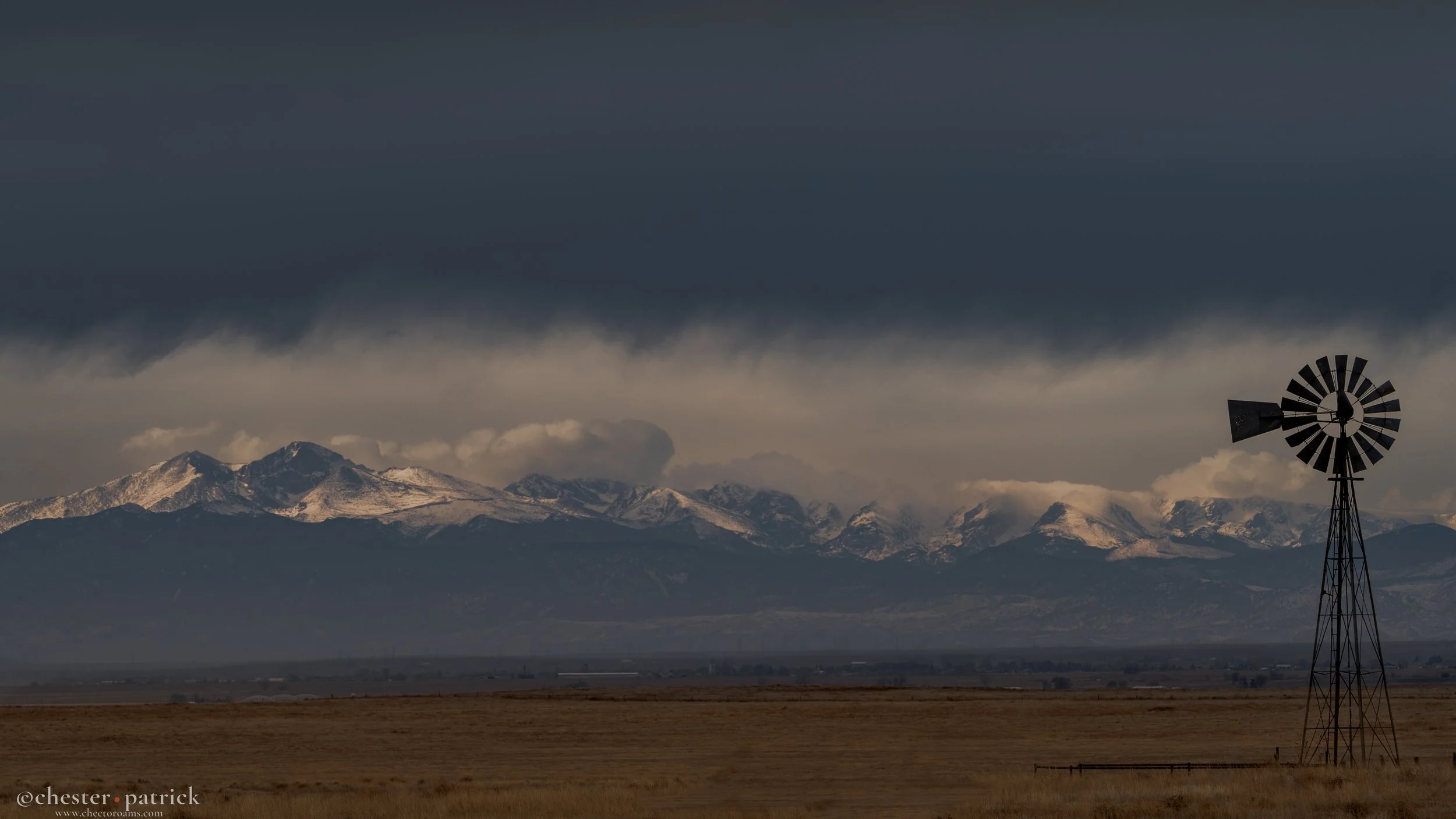 Snow-capped mountains under dark, cloudy sky with a windmill in the foreground