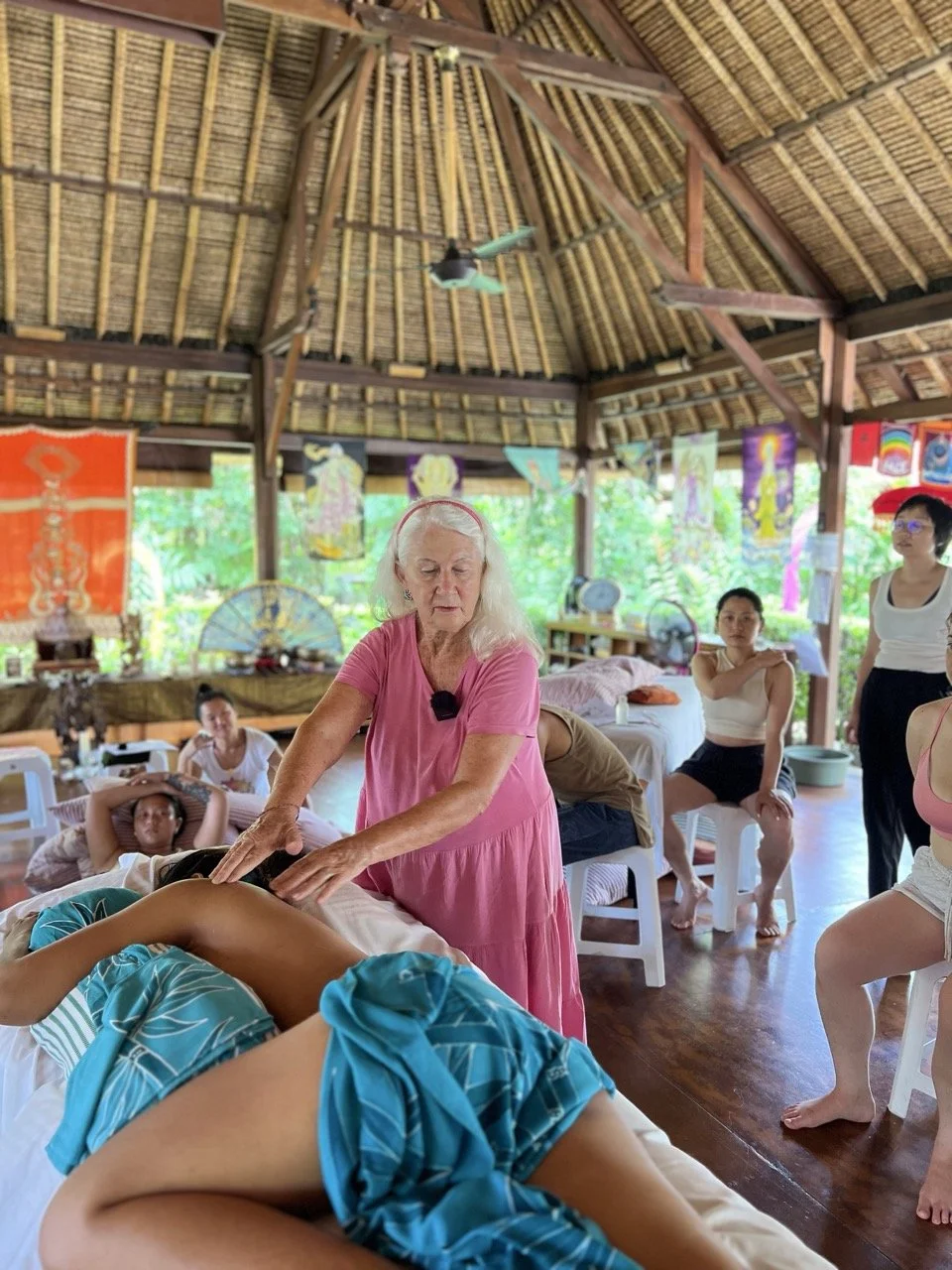 A woman receiving a massage at a workshop, surrounded by an audience in a rustic, bright space decorated with artwork.