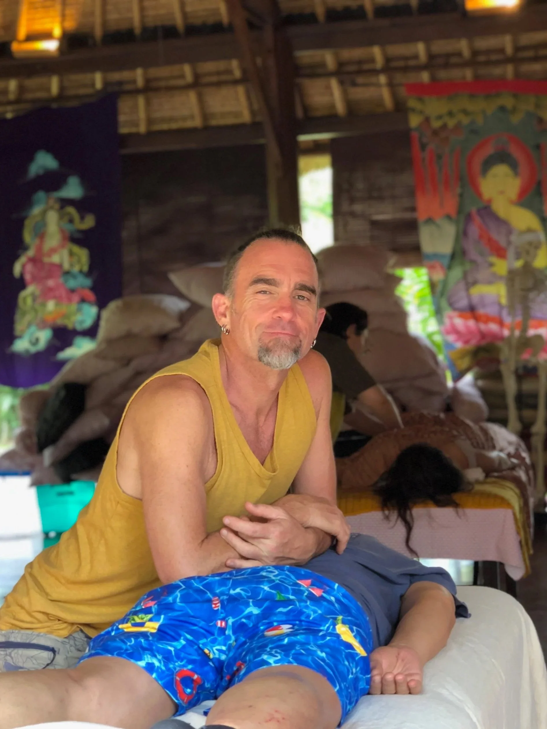 A group of people gathered around a person lying on a massage table inside a hut with a thatched roof, participating in a traditional healing or spiritual ceremony.