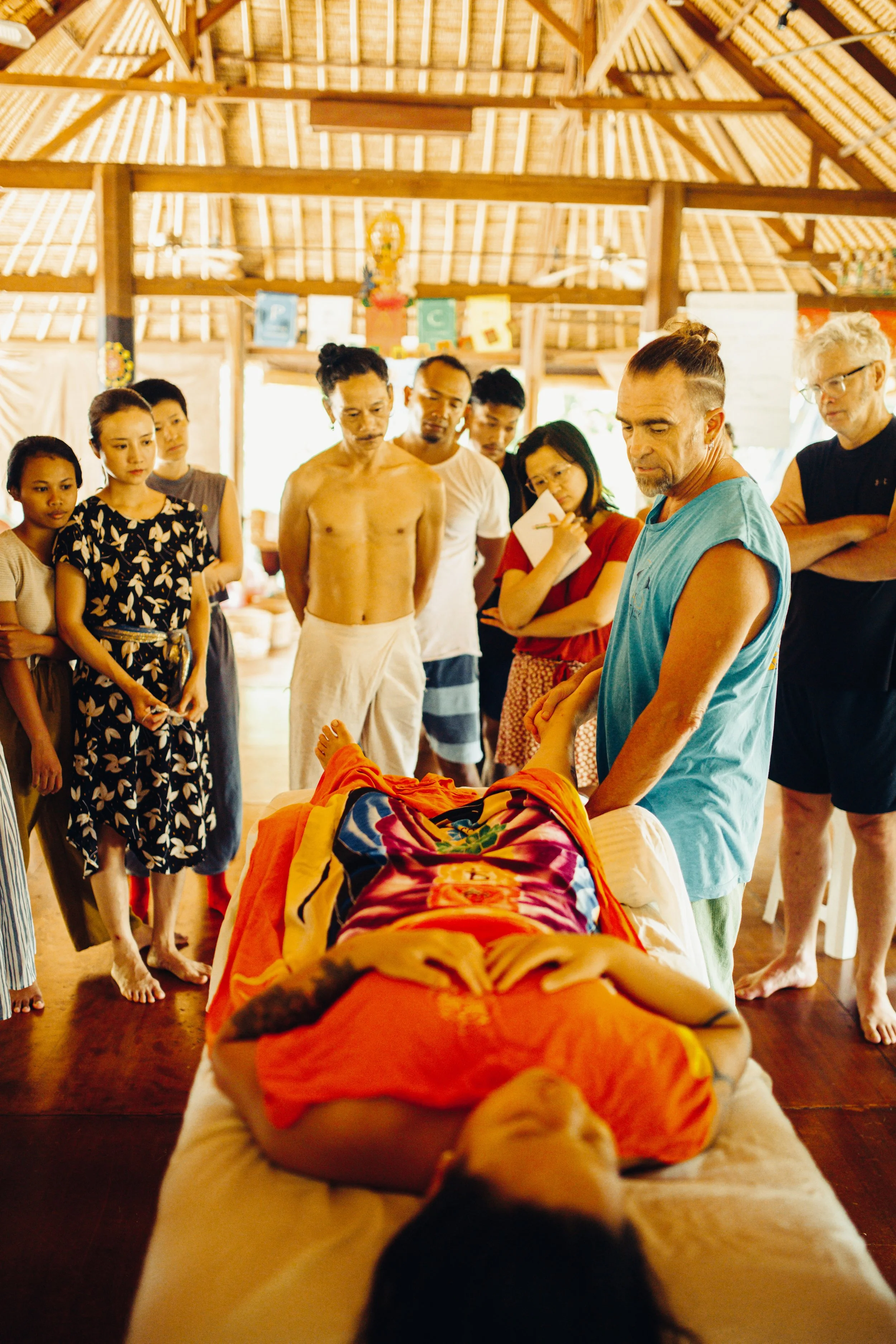 A group of people gathered around a person lying on a massage table inside a hut with a thatched roof, participating in a traditional healing or spiritual ceremony.