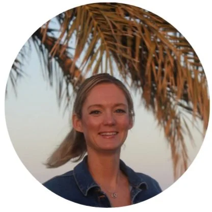 Jenny smiling outdoors, standing in front of a palm tree with brown fronds, during daytime.