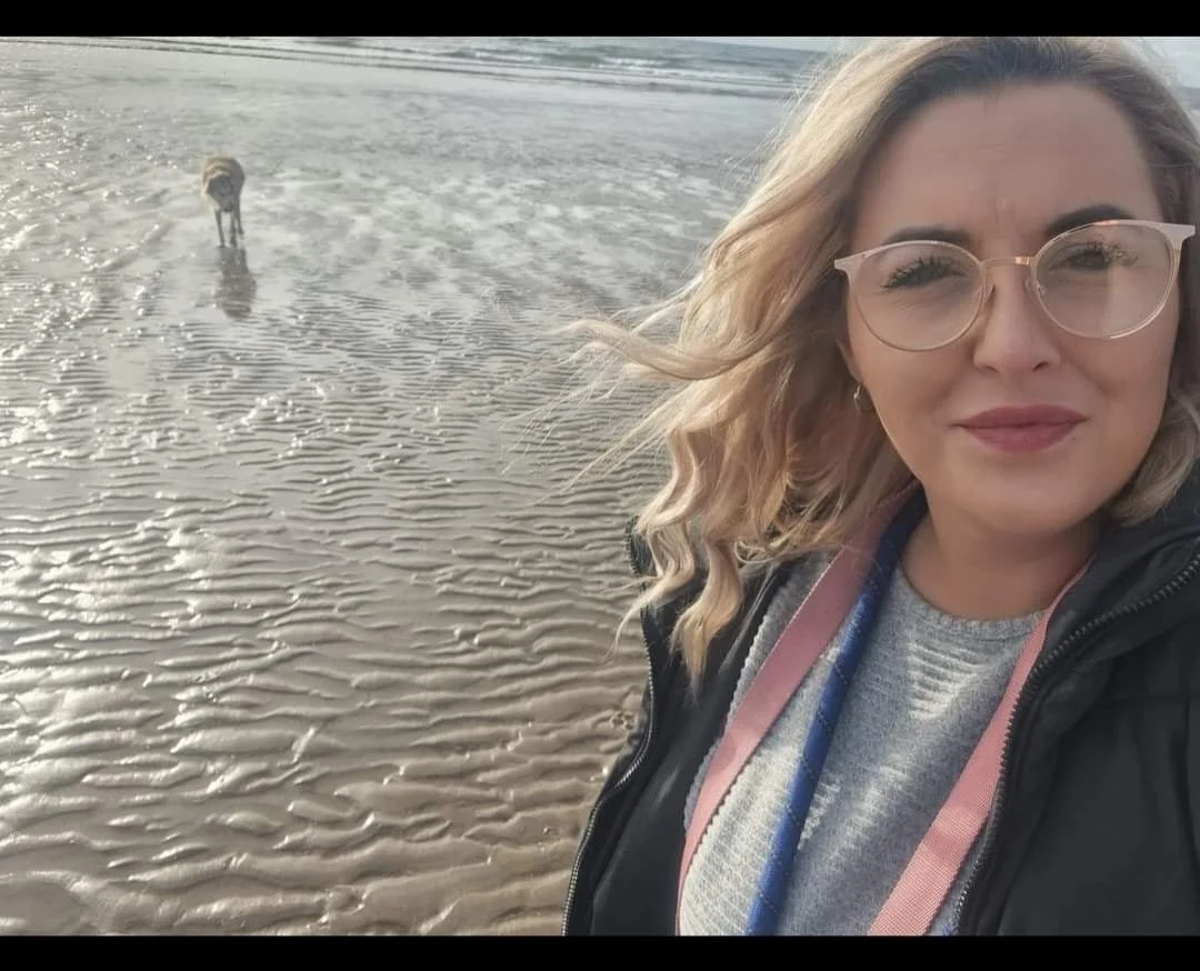 Jayne helps neurodivergent and chronically ill folks with admin and oraganisation. She has shoulder-length blonde hair and is wearing gold-rimmed glasses and a grey jumper with a navy coat. She is walking her dog on the beach.