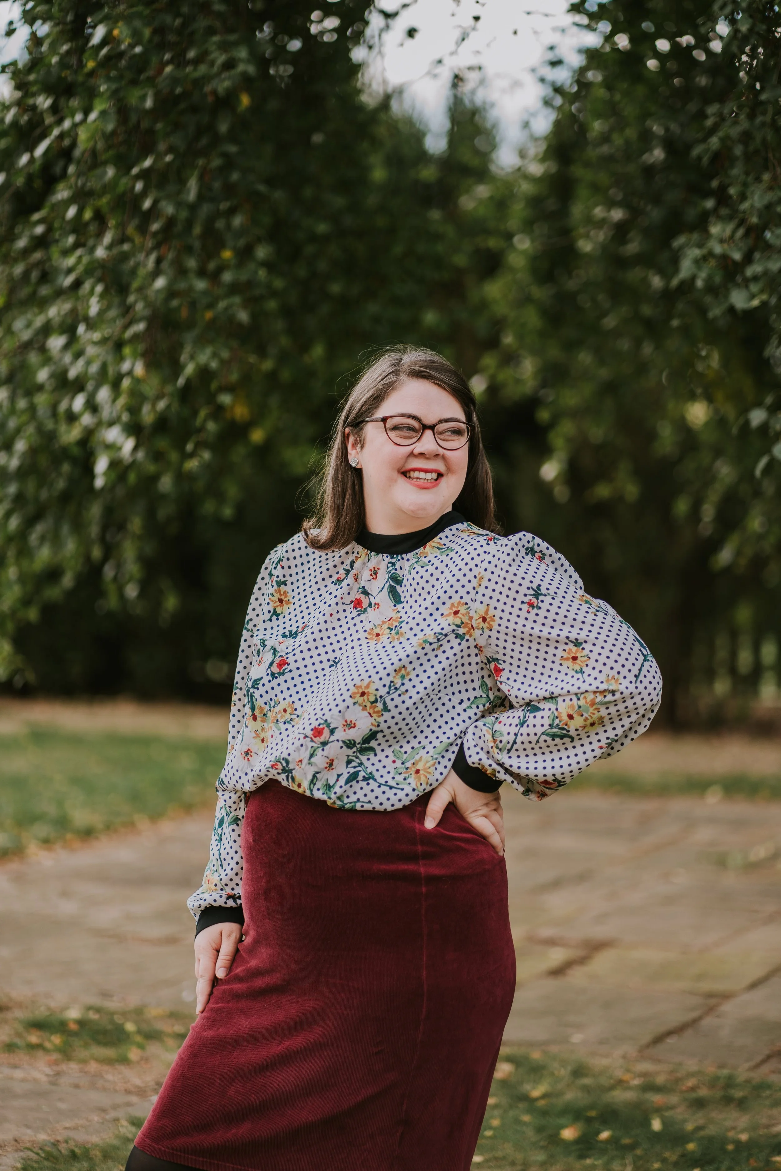Laura supports you apply for Access to Work ADHD or autism coaching and is shown here outdoors in a polka dot floral blouse with black glasses and shoulder-length hair smiling, one hand on her hip.