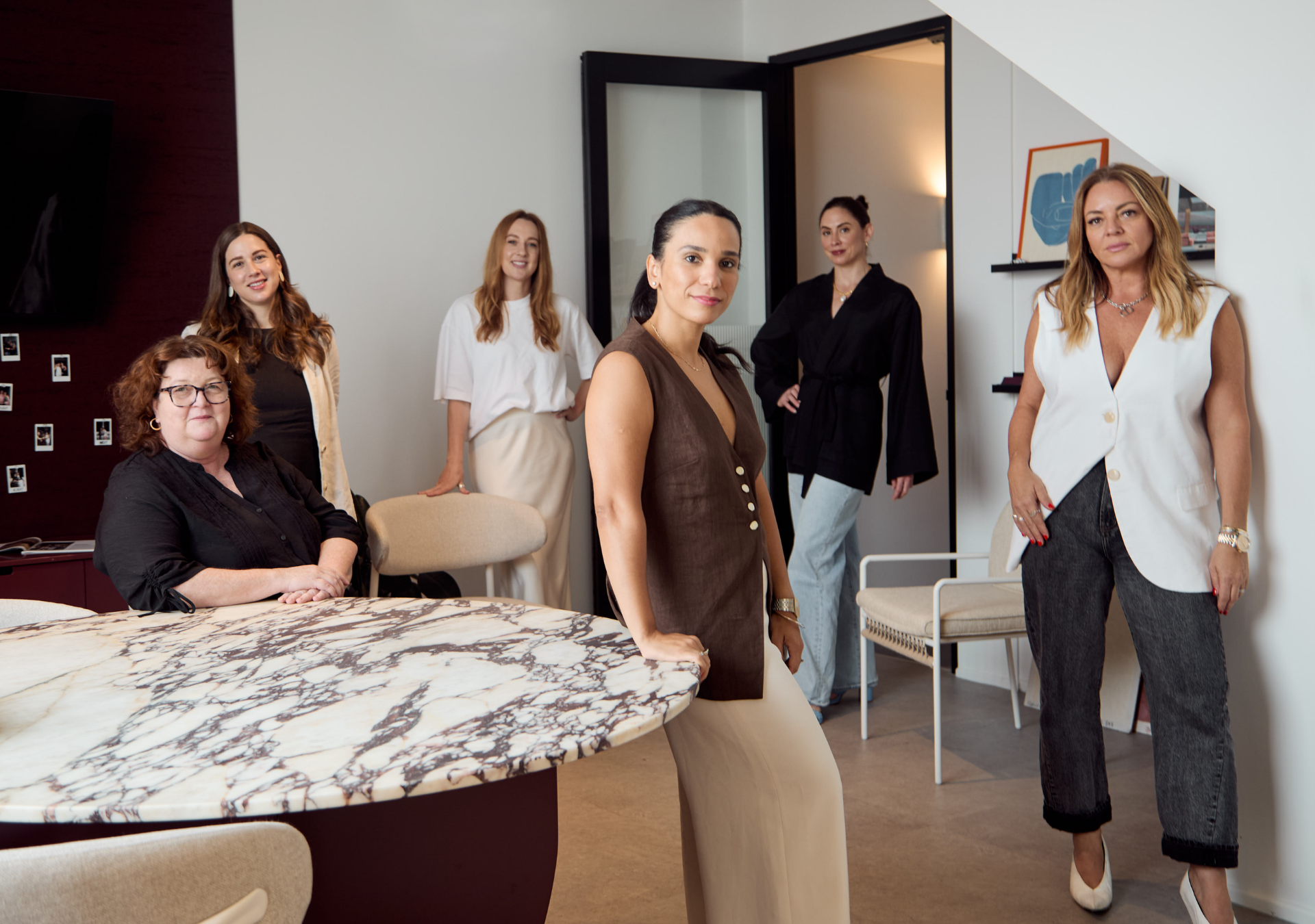 Six women in an office or conference room, standing and sitting, with a marble table and modern decor in the background.