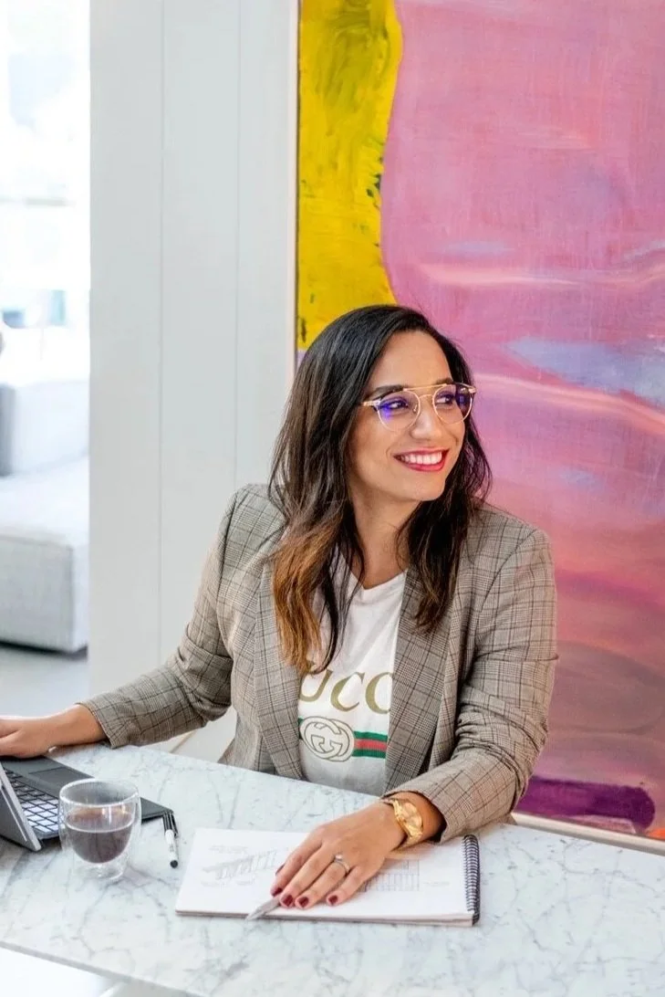 A woman with glasses and a checkered blazer sitting at a white marble table, smiling, with a laptop, a glass of water, and some documents in front of her.