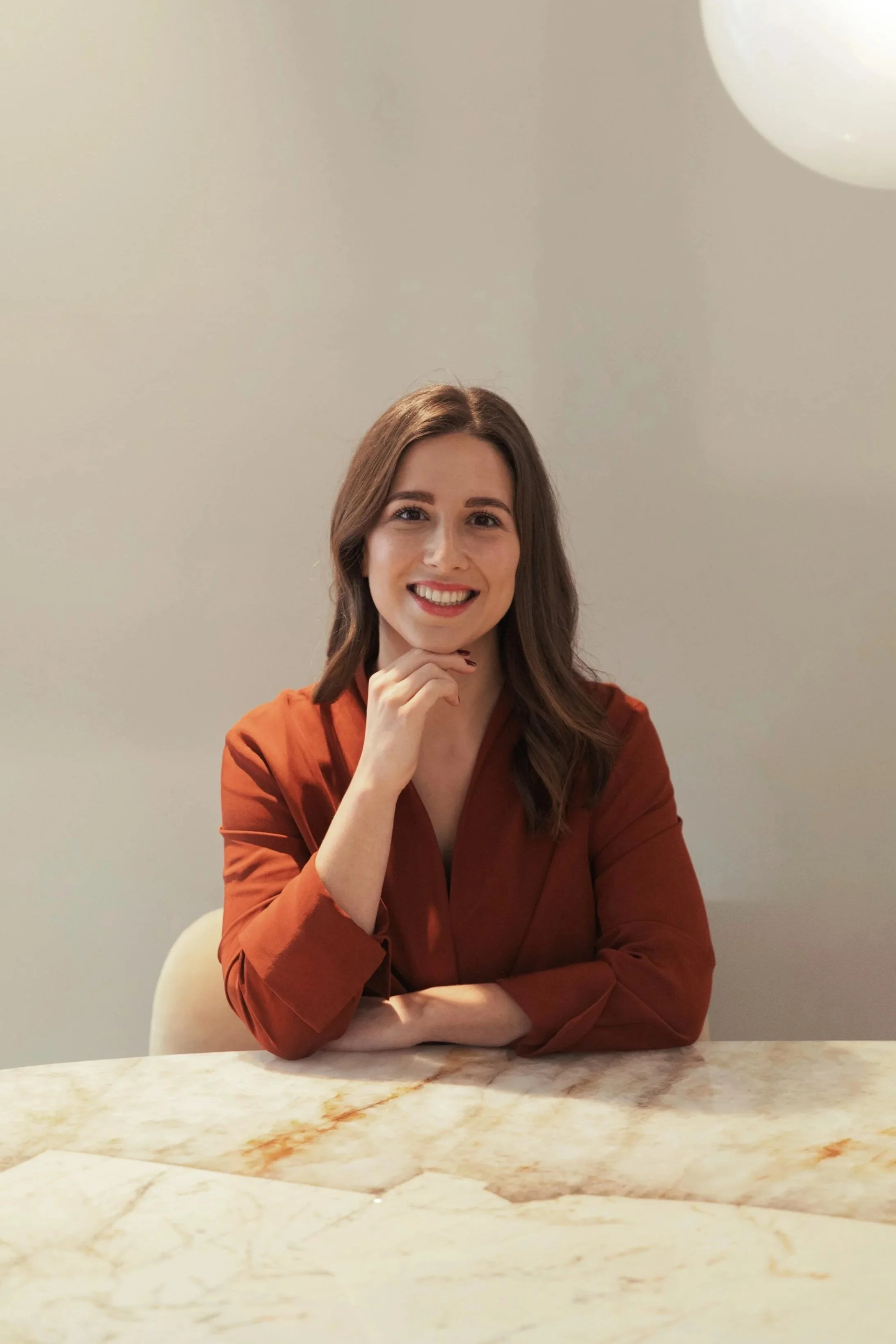 A woman with long brown hair and a warm smile sitting at a marble table, wearing a rust-colored blouse, with her hand resting on her chin, in a minimalistic room with a white wall and circular light fixture ceiling.
