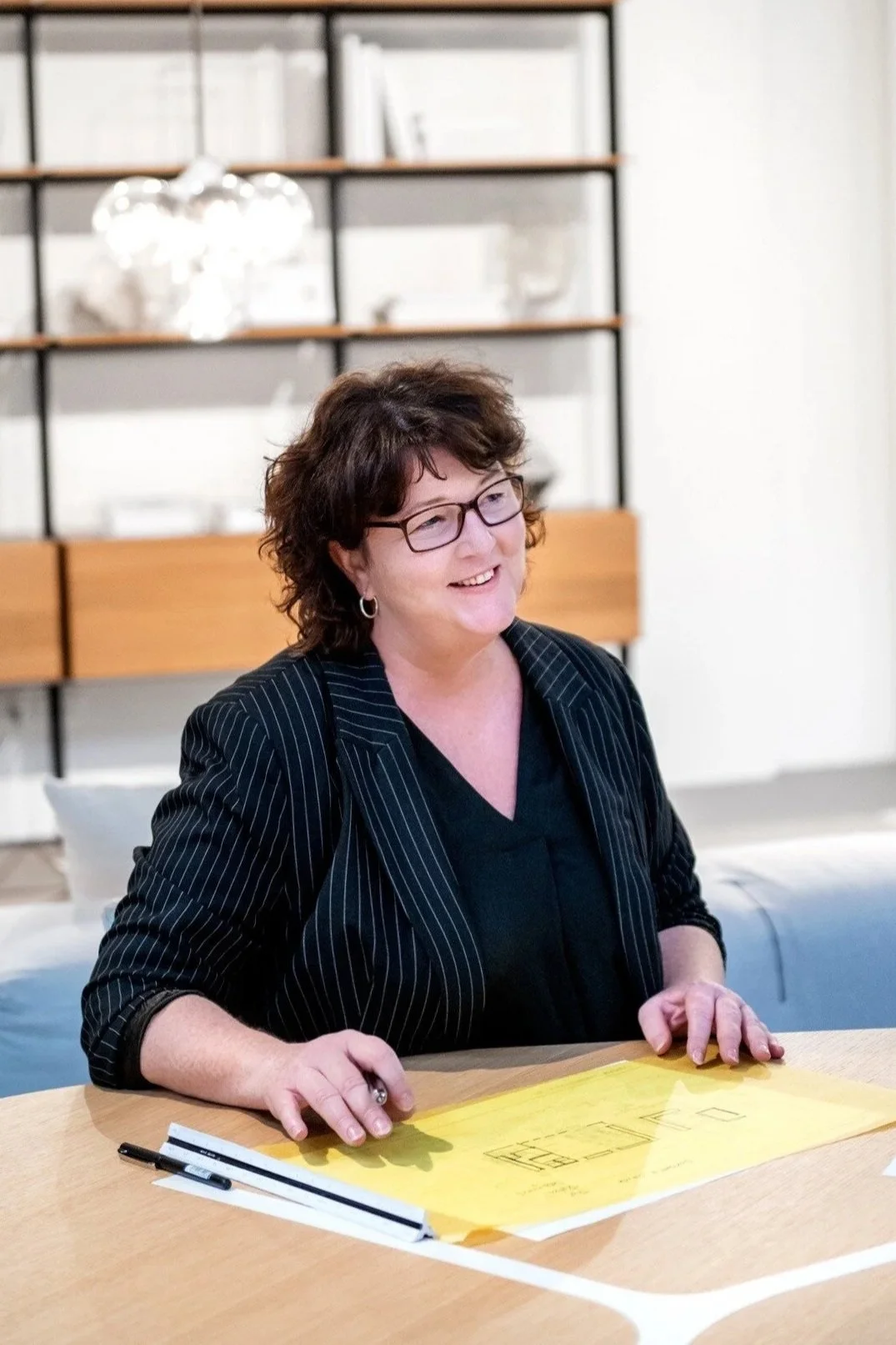 A woman with glasses and short curly hair sitting at a table with yellow paper and black markers, smiling indoors with a shelving unit and white wall in the background.