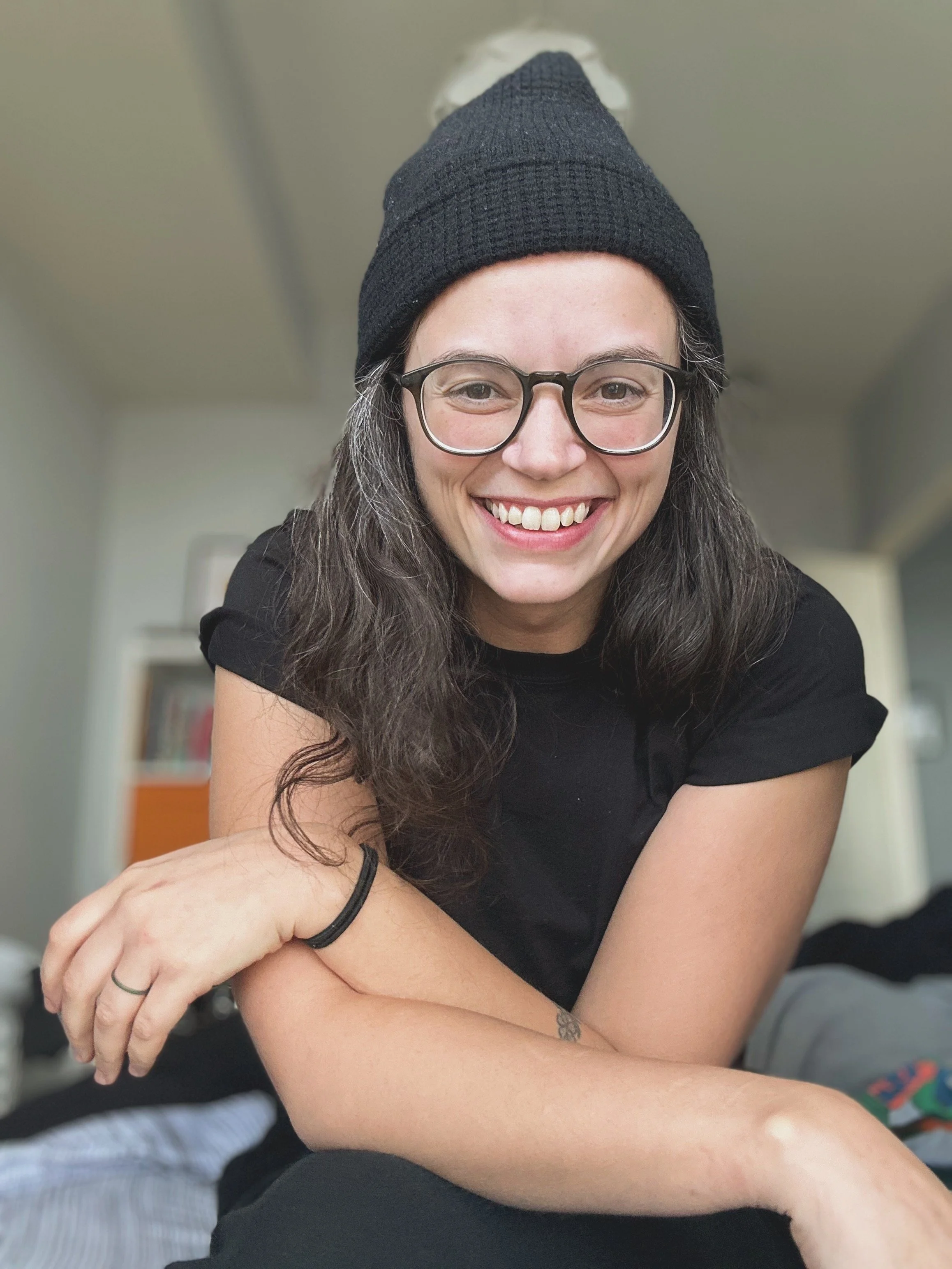 A young woman with glasses, wearing a black beanie and black t-shirt, smiling and leaning forward in a relaxed pose indoors.