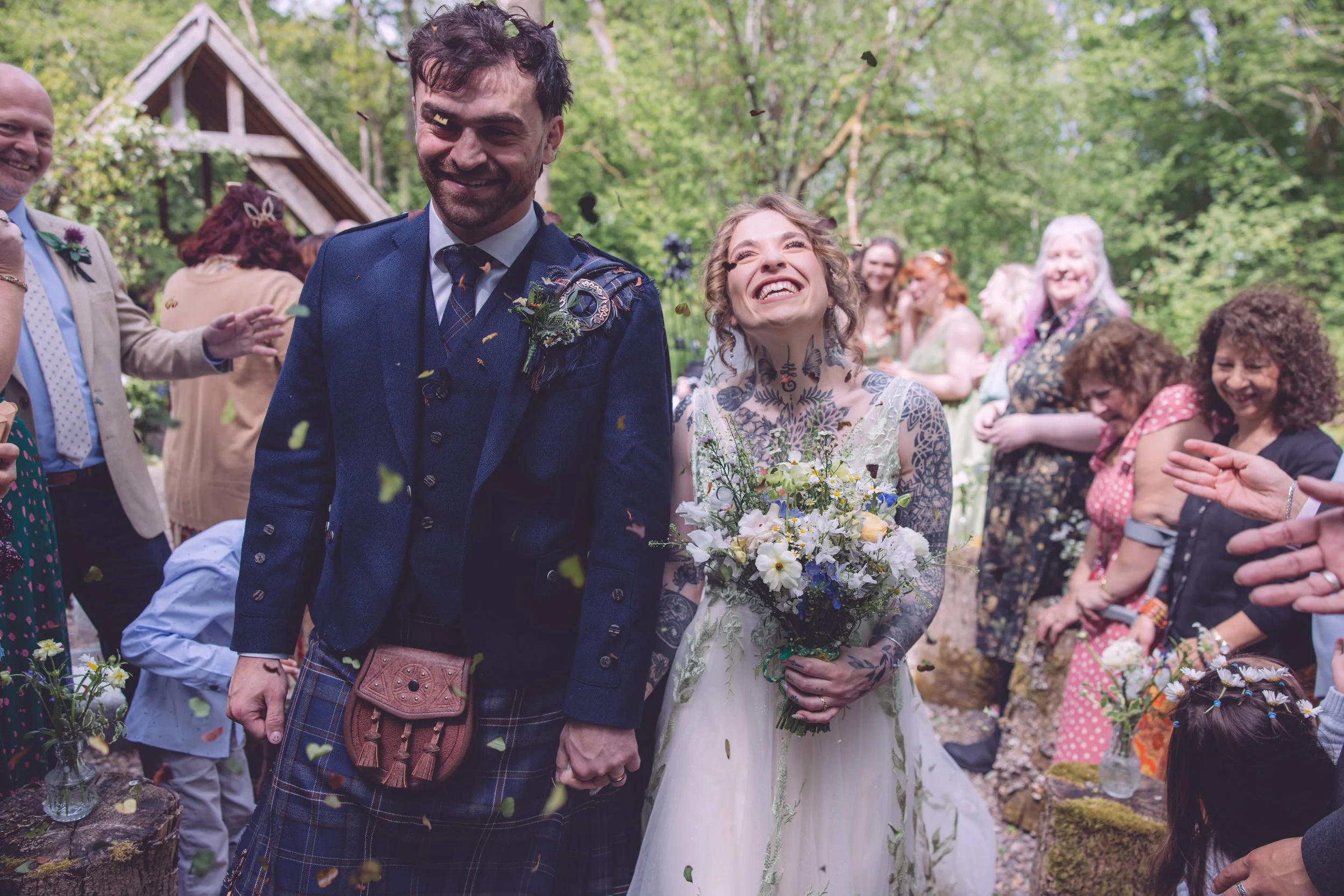 A smiling bride and groom holding hands during their outdoor wedding celebration surrounded by family and friends in a wooded area, with confetti falling.