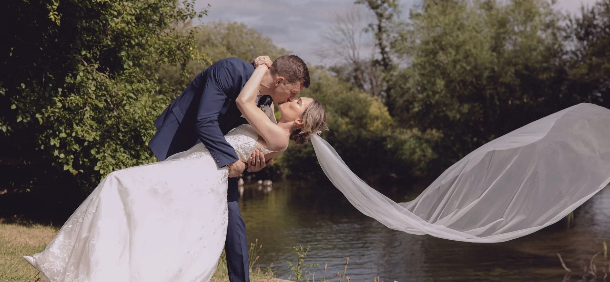 Groom dipping his bride by the water, veil flowing
