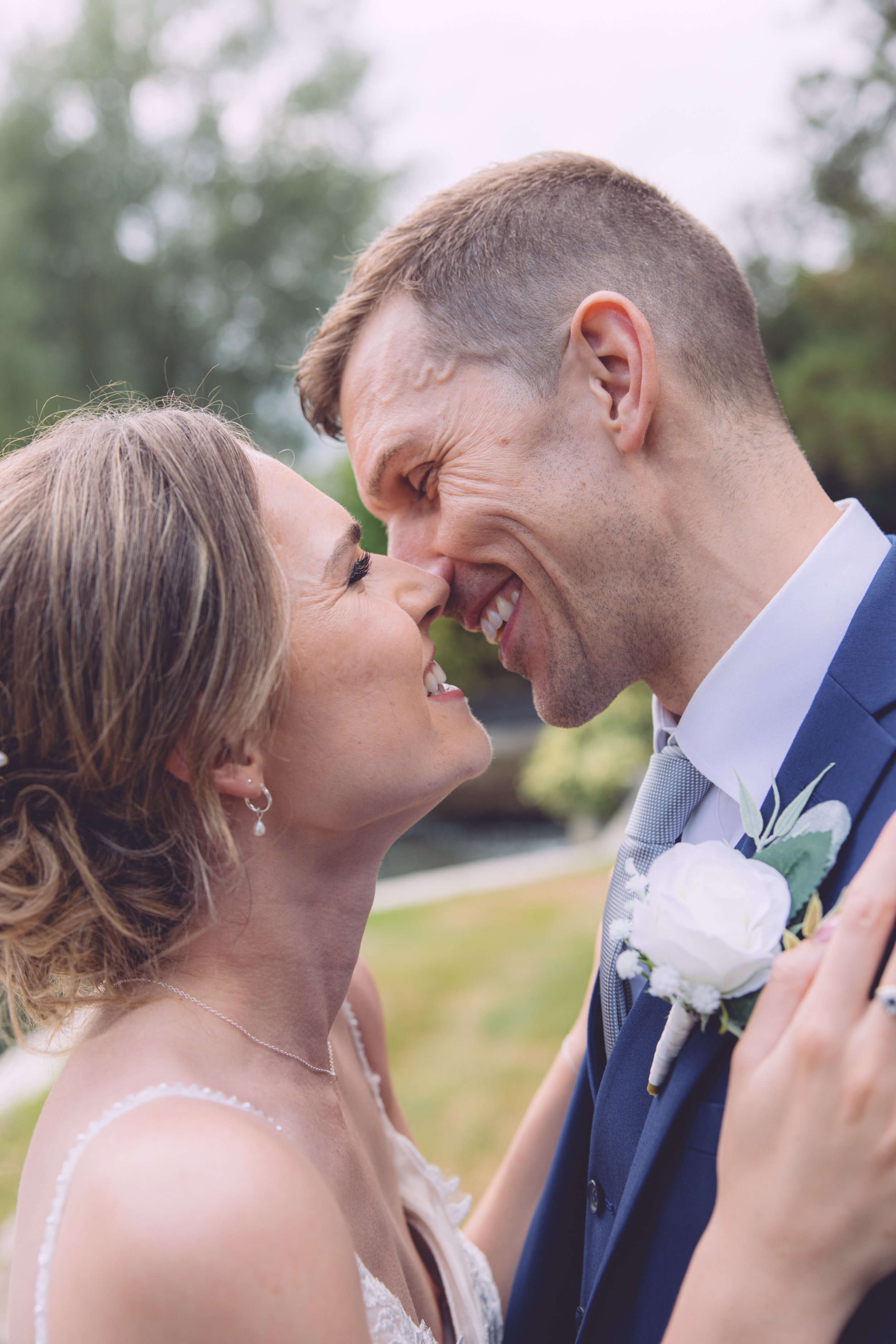 A bride and groom are smiling closely together, touching foreheads in a joyful moment outdoors, with green trees in the background.