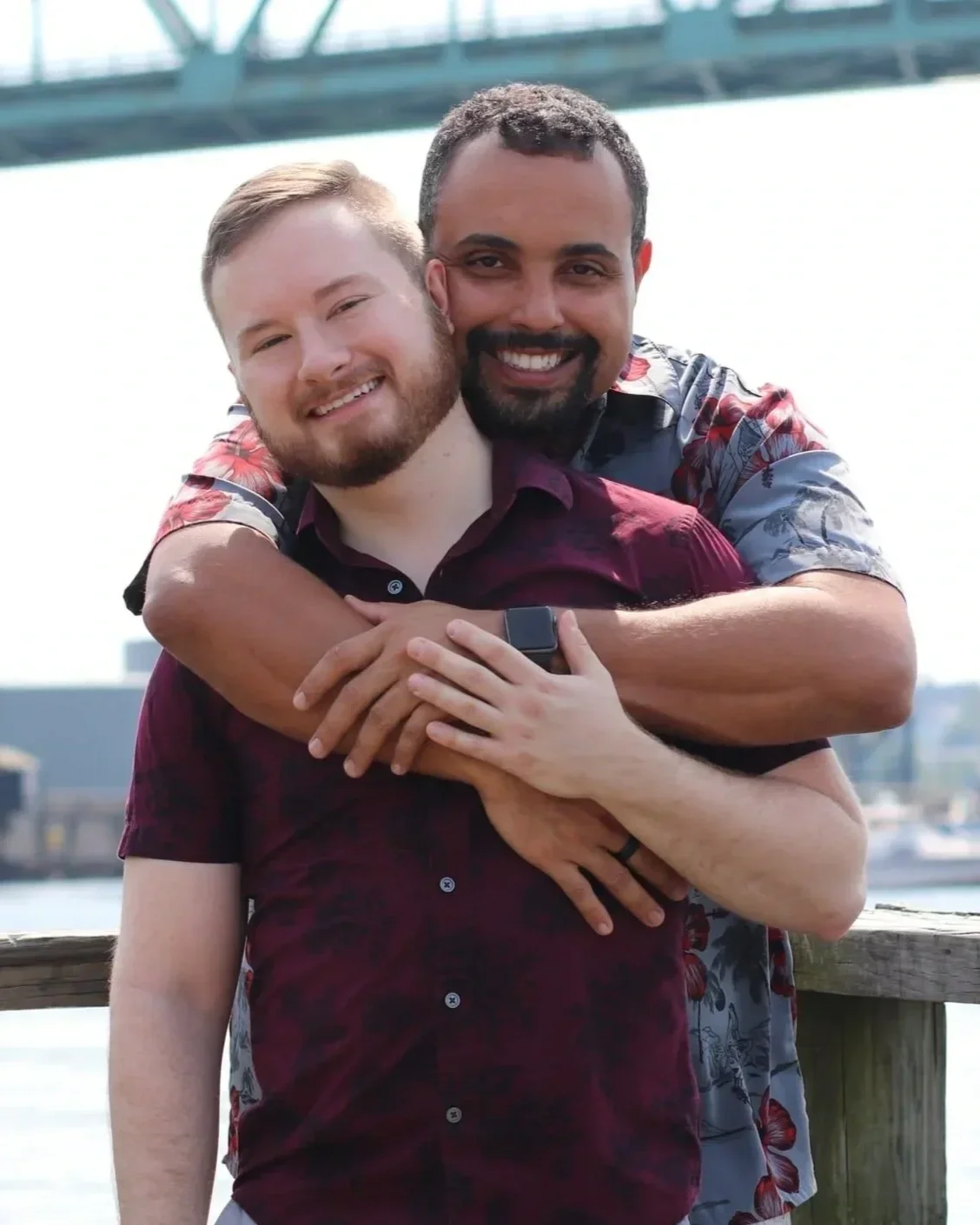 Two men pose together facing the camera, with one man standing behind the other and hugging him from behind. One man has lighter skin and the other has medium-brown skin, and both appear relaxed and comfortable.