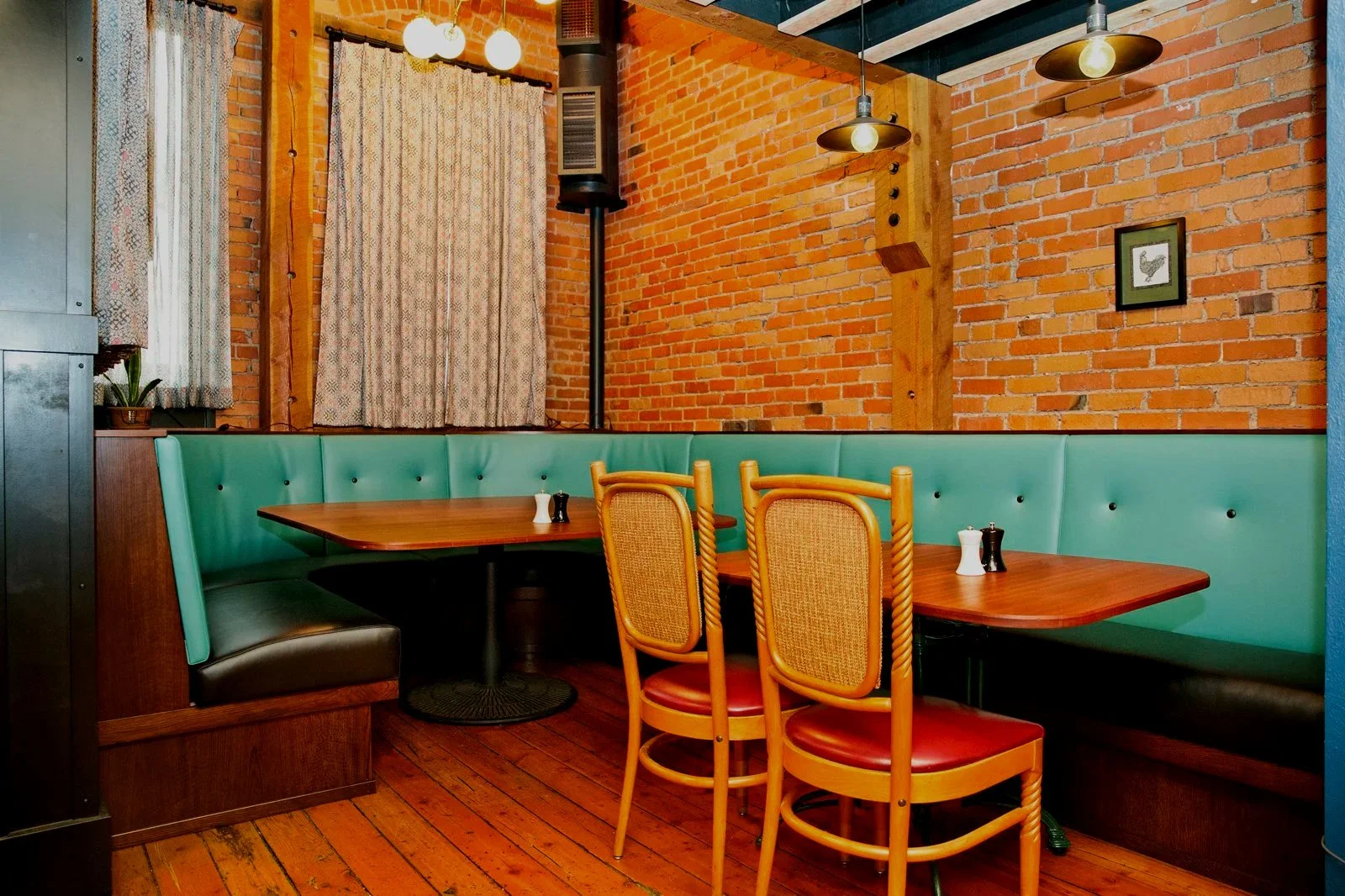 Inside a cozy restaurant with brick walls, a large green booth, a wooden table, and wooden chairs with woven backs. There are curtains on the window, a small potted plant, framed artwork on the wall, and hanging light fixtures.
