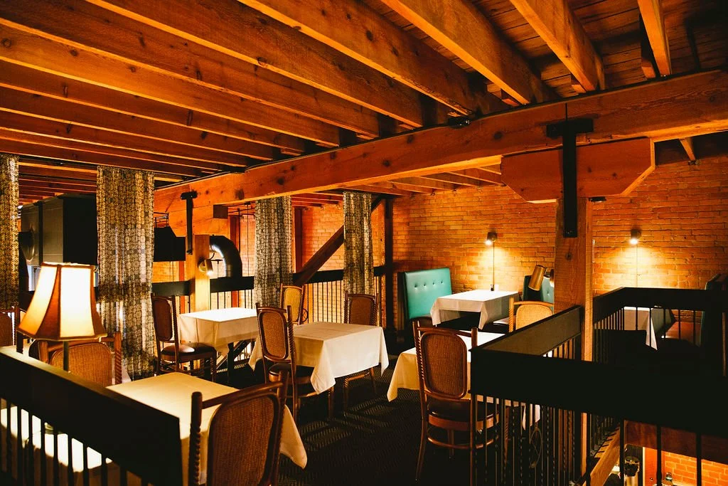 Interior of a cozy restaurant with brick walls, wooden beams, chairs, and warm lighting.