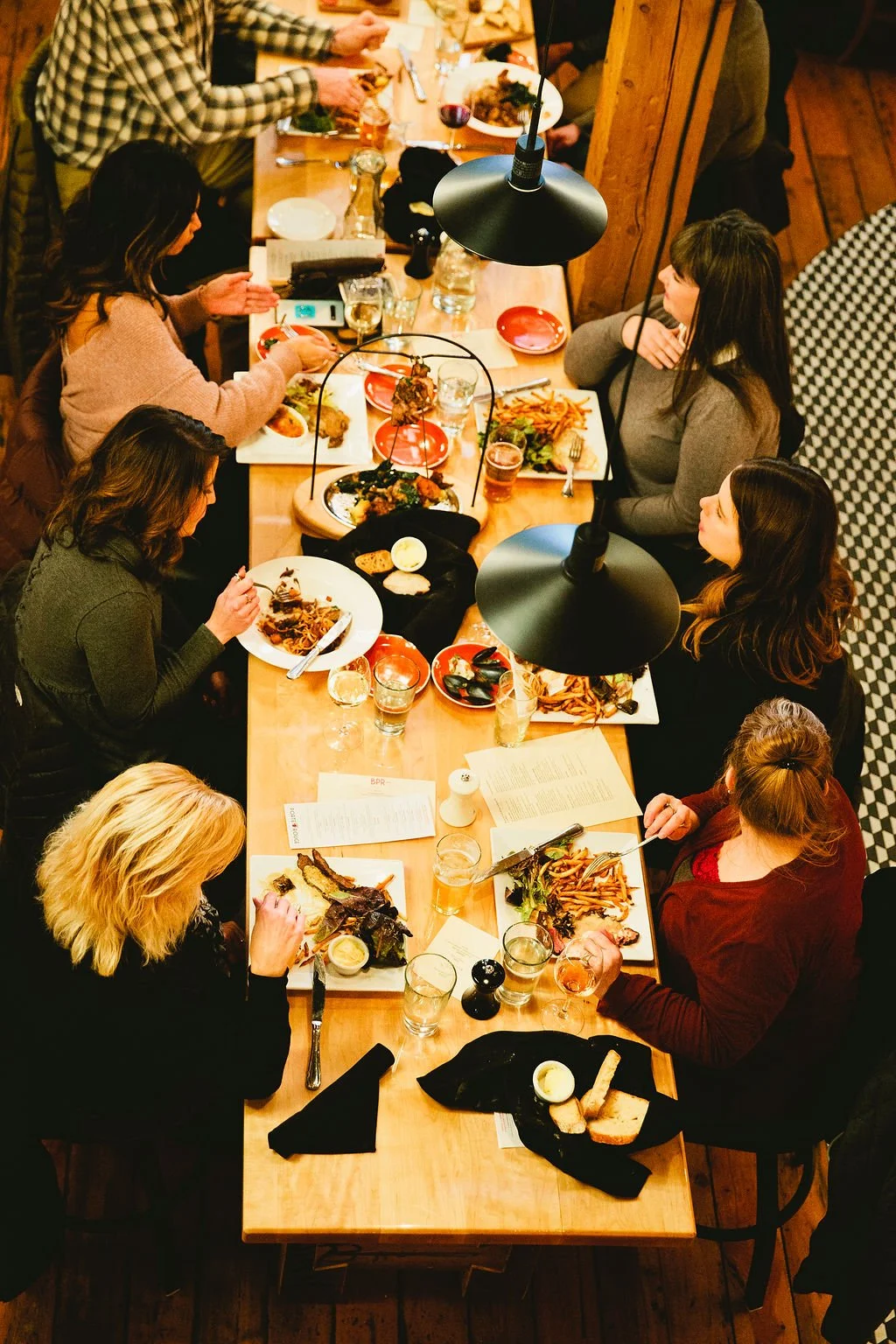 A group of women seated around a dining table enjoying a meal together at a restaurant, with various dishes, drinks, and utensils on the table.