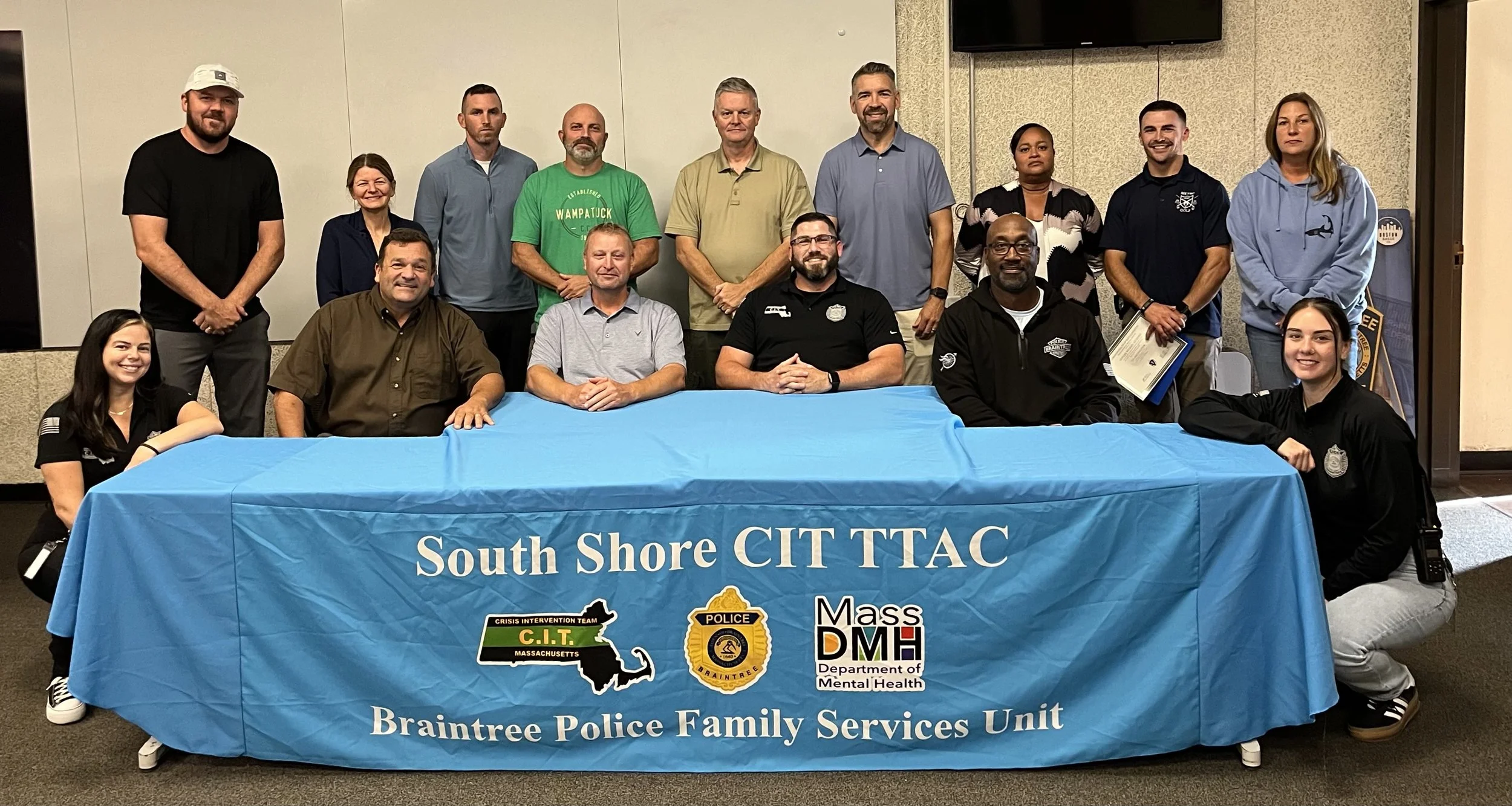 A group of people, including police officers and community members, gathered behind a blue tablecloth with the text 'South Shore CIT TTAC,' representing the Braintree Police Family Services Unit. They are indoors against a beige wall, smiling, and dr