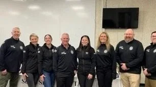 Seven individuals standing together indoors, wearing black jackets or sweatshirts with logos, in front of a wall with a mounted TV.