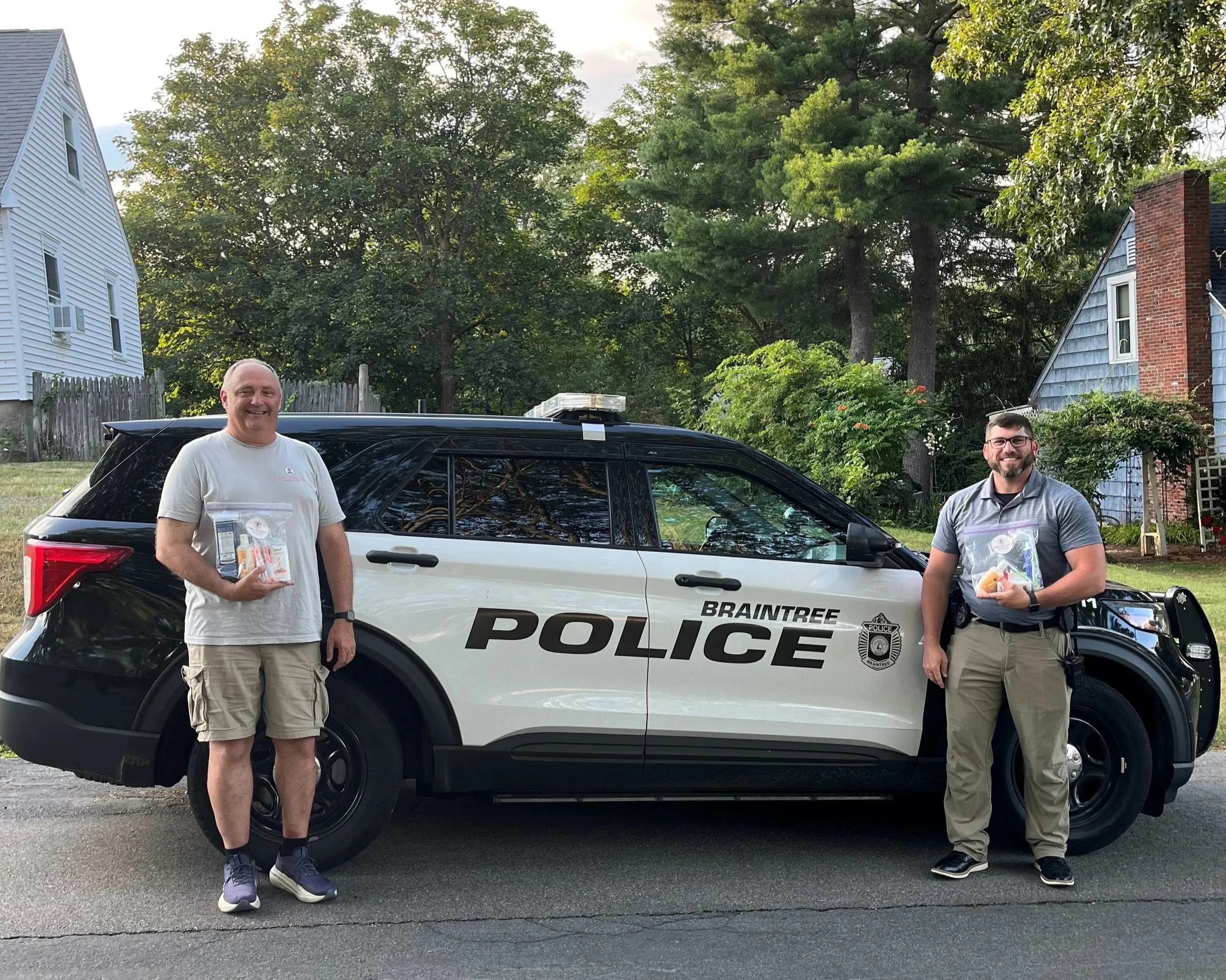 Two men standing in front of a police SUV, smiling, holding bags of snacks, with trees and houses in the background.
