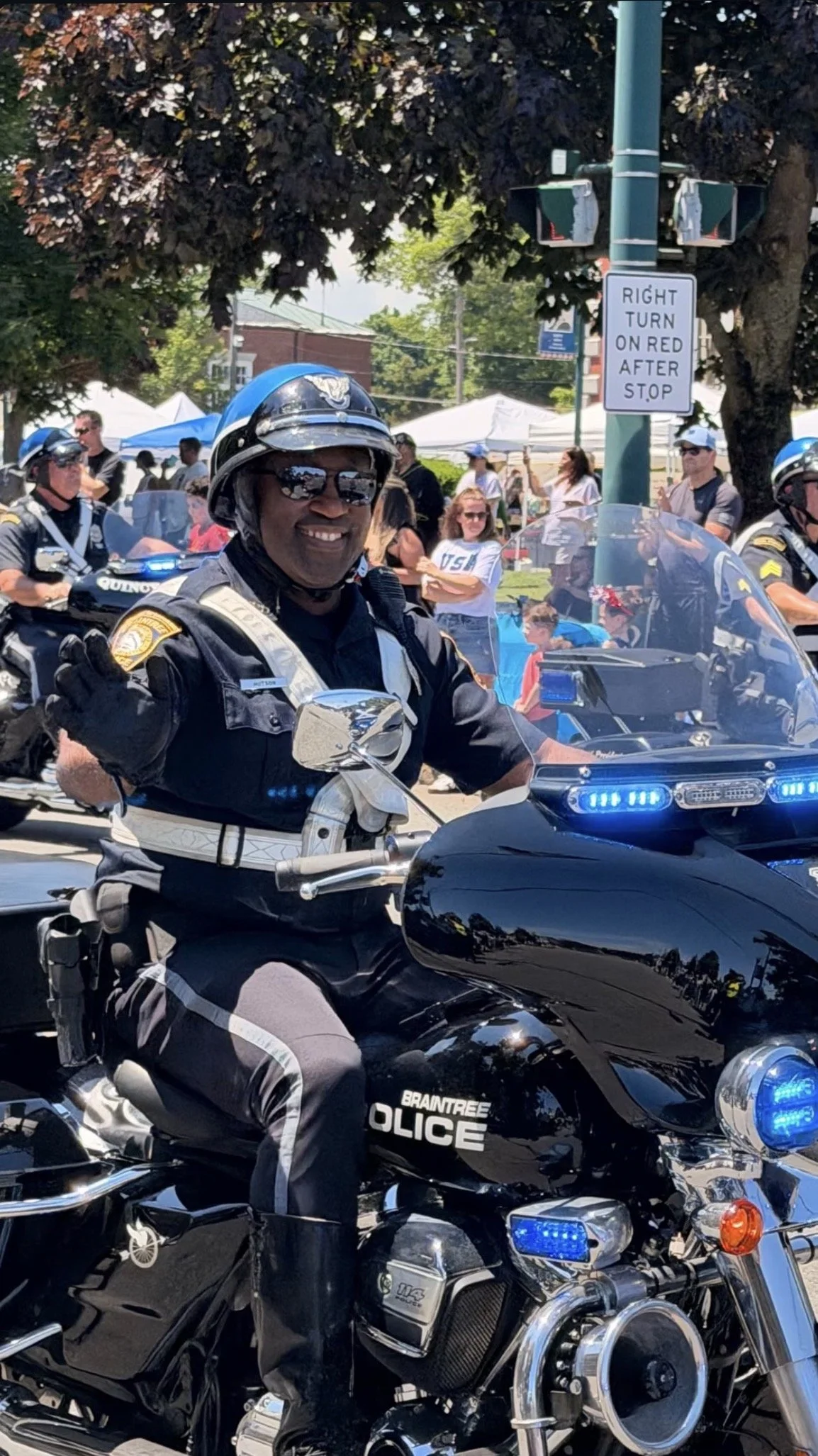 A man wearing sunglasses, police officer motorcycle uniform and a name tag that says 'C. Hutson' waving while riding a police motorcycle during a Braintree parade in the summertime..