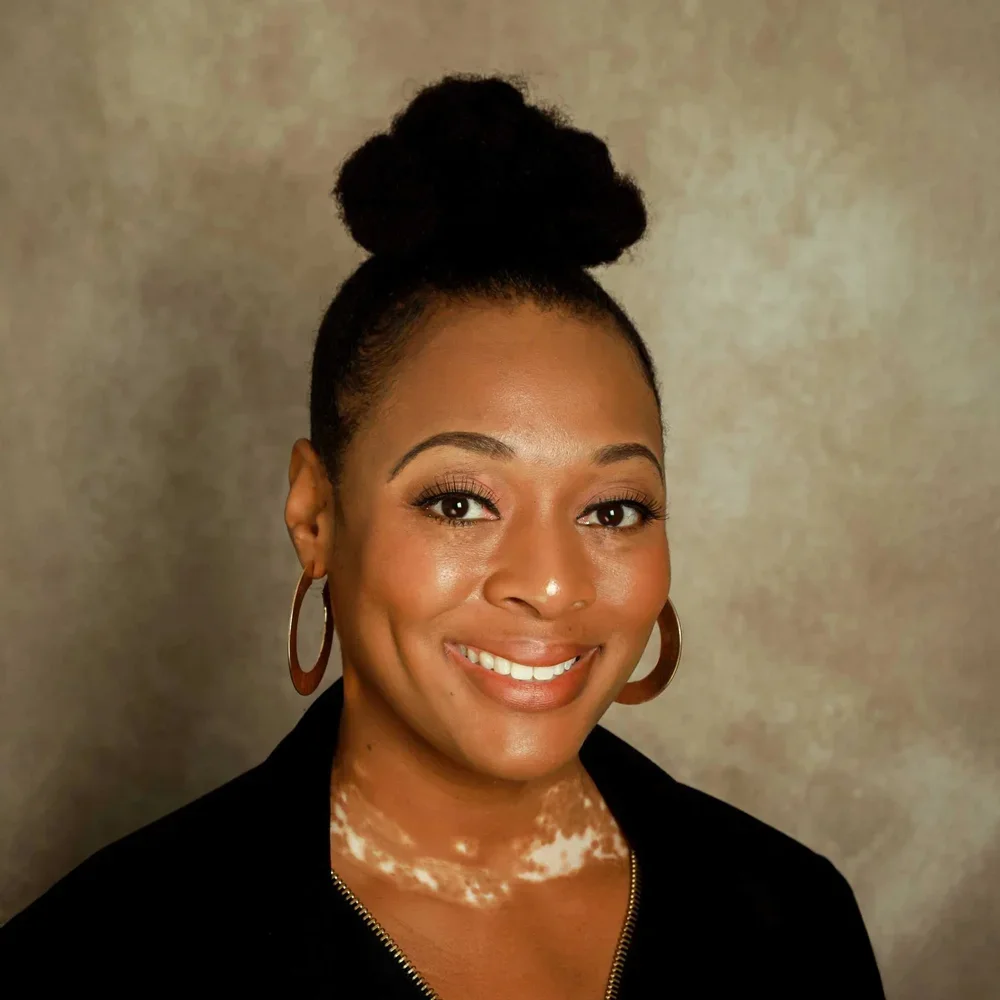 Portrait of a woman with dark hair styled in an updo, wearing large hoop earrings and a black top, smiling in front of a neutral background.