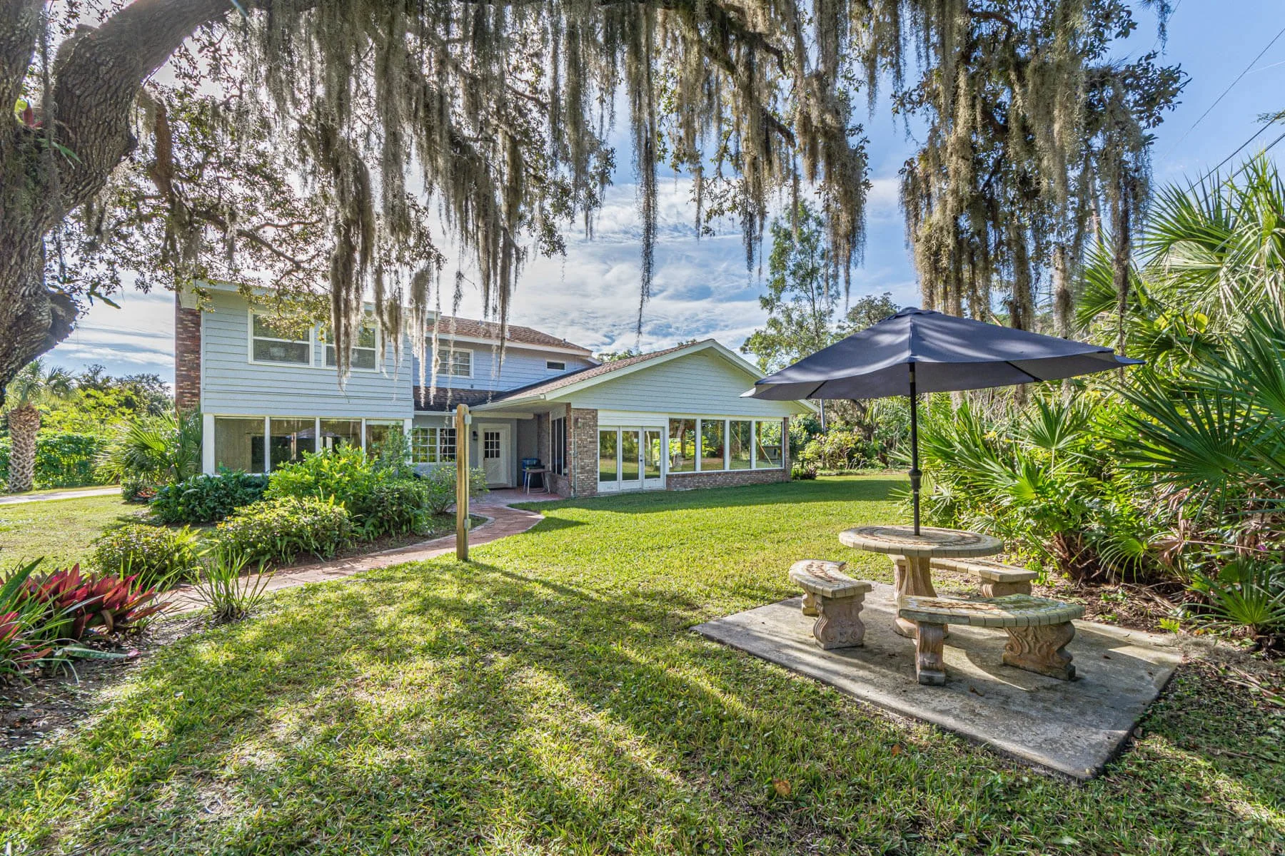 A house with large windows and a brick patio in the backyard, shaded by trees with hanging moss and an umbrella over a stone picnic table.