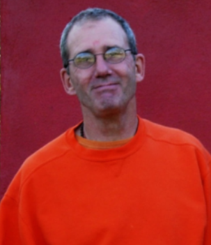 A man with short hair and glasses, wearing an orange shirt, standing in front of a red background.