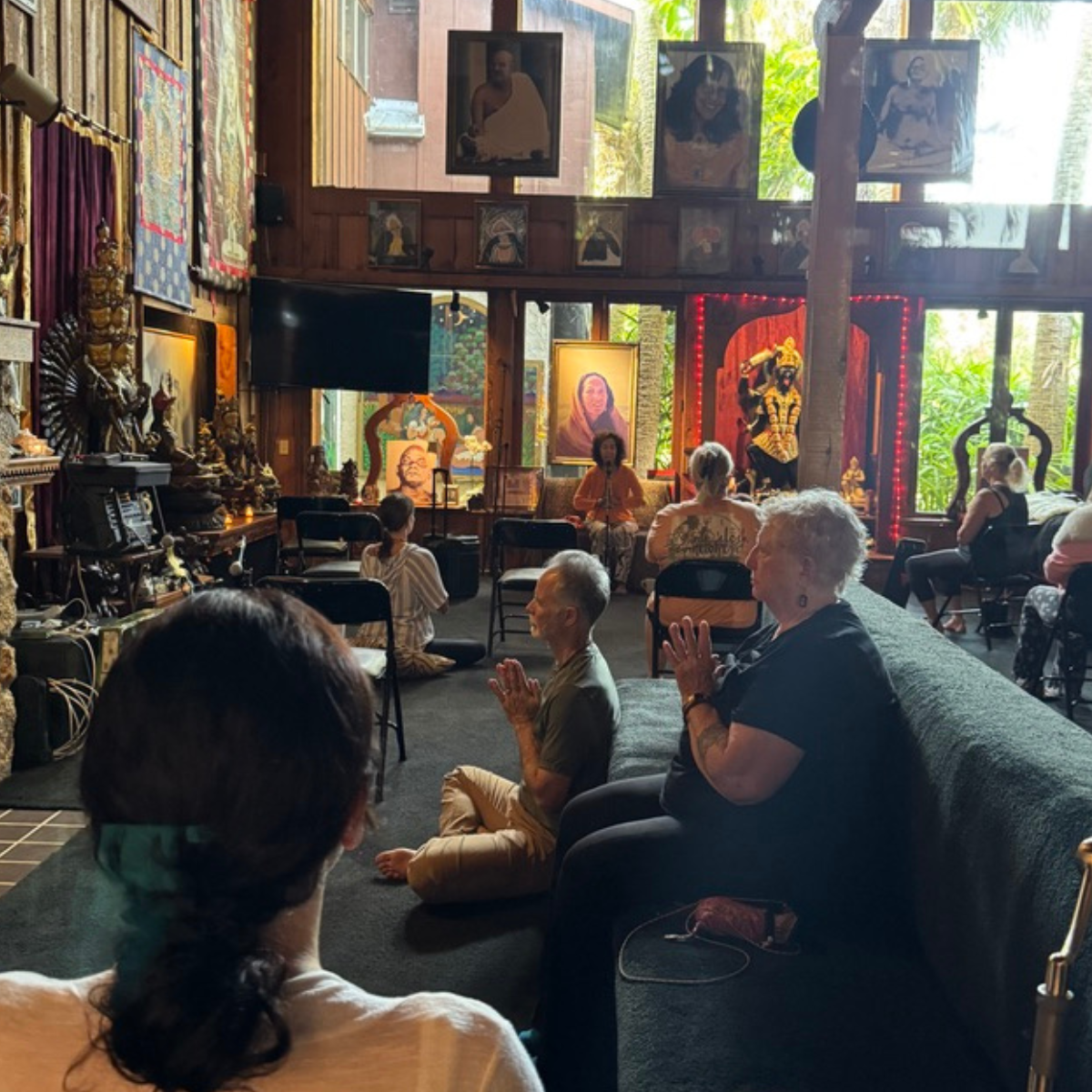 People participating in a meditation or prayer session in a room decorated with spiritual and religious art, sculptures, and portraits, with a woman leading the session seated on a chair at the front.