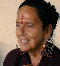 Close-up of an elderly woman with traditional face markings, wearing large hoop earrings and a dark top.