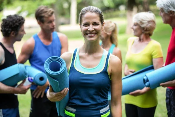 Woman smiling while holding a yoga mat, surrounded by other people outdoors.