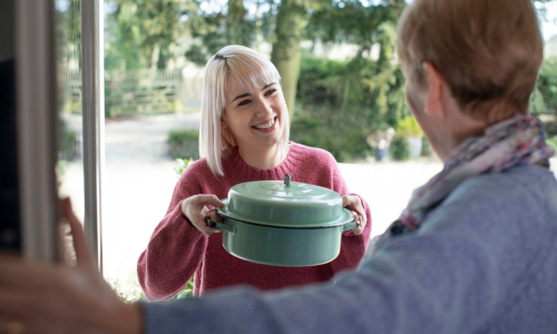 A woman smiling and handing a green casserole dish to an older man at the doorway of a house, with outdoor greenery in the background.