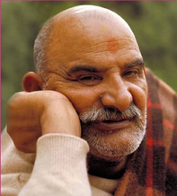 Close-up of an elderly man with a bald head, white beard, and mustache, resting his chin on his hand and smiling softly.