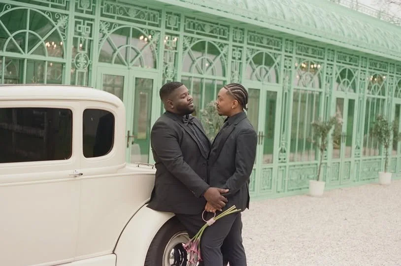 Two people dressed in black suits sharing an intimate moment outside a green ornate building, with one holding a small bouquet of flowers.