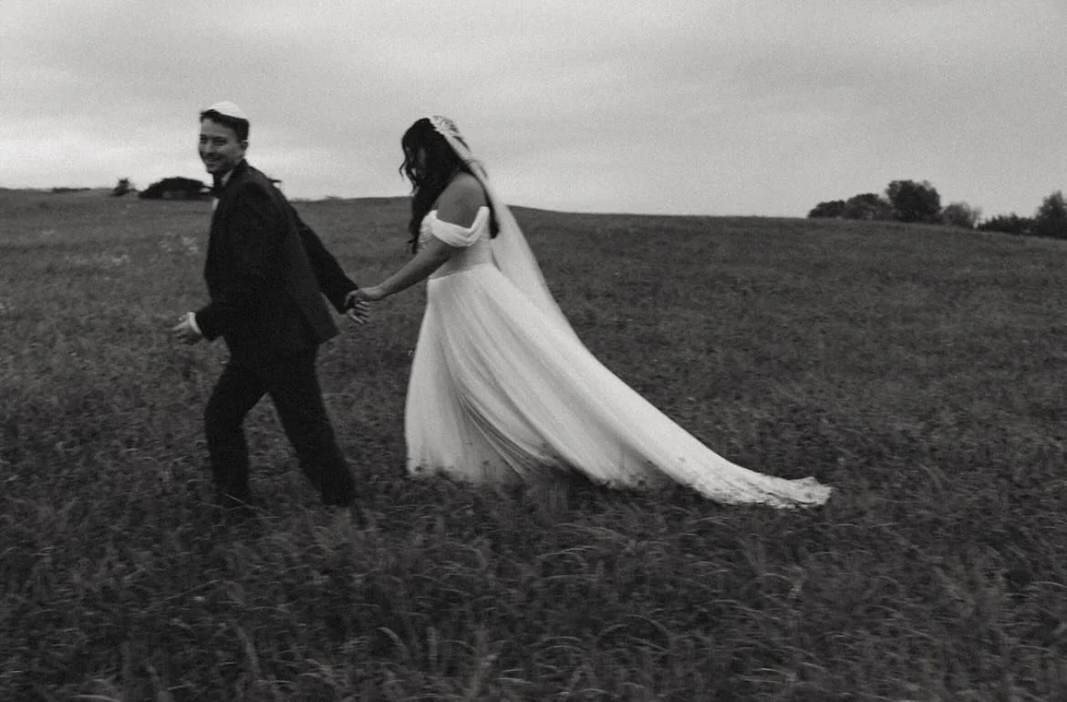 A black-and-white photo of a bride and groom walking through a grassy field, holding hands. The bride is wearing a long, flowing wedding dress with a veil, and the groom is in a suit with a backpack.