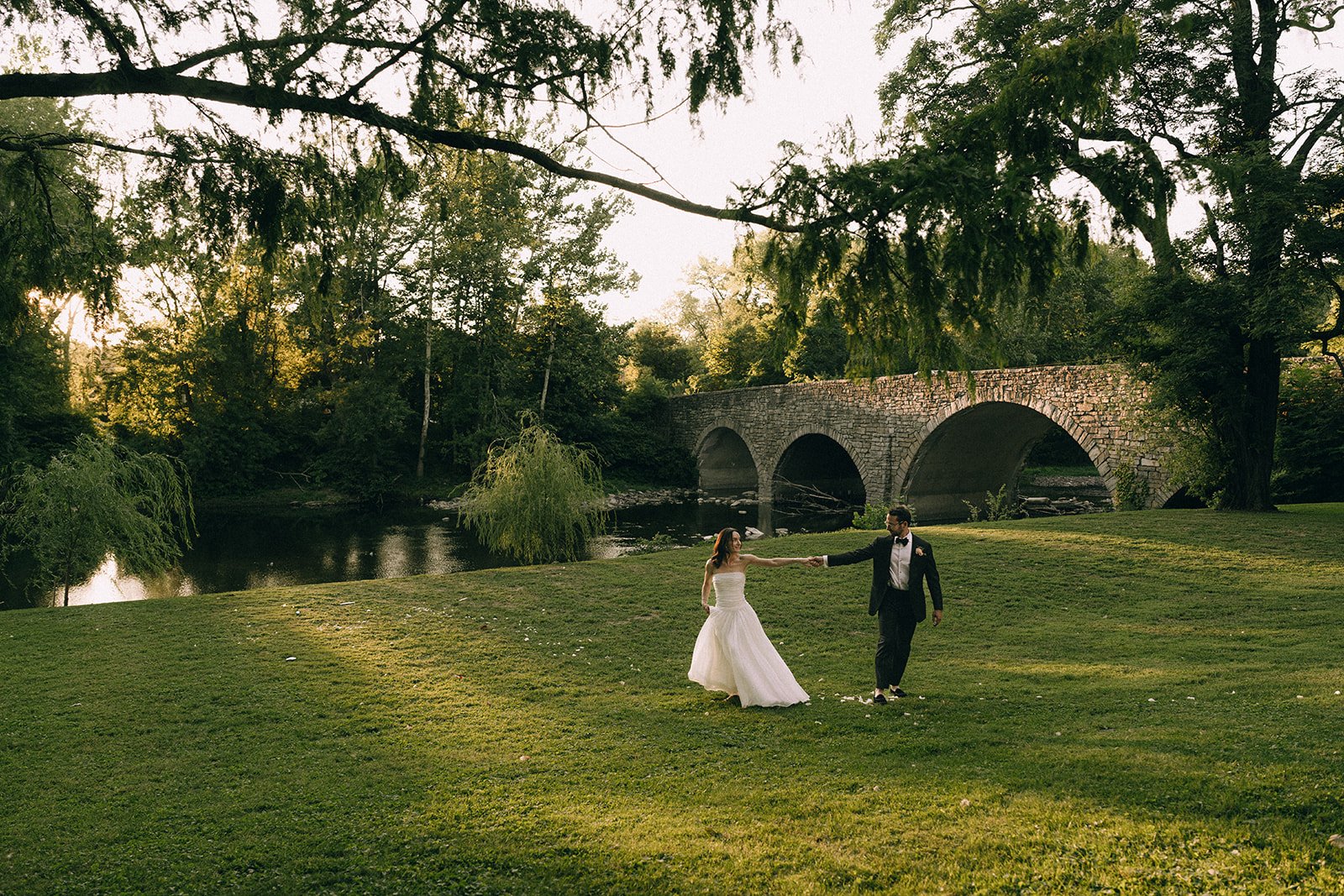 A bride and groom dancing on a grassy area near a river, with a stone bridge and trees in the background during sunset.