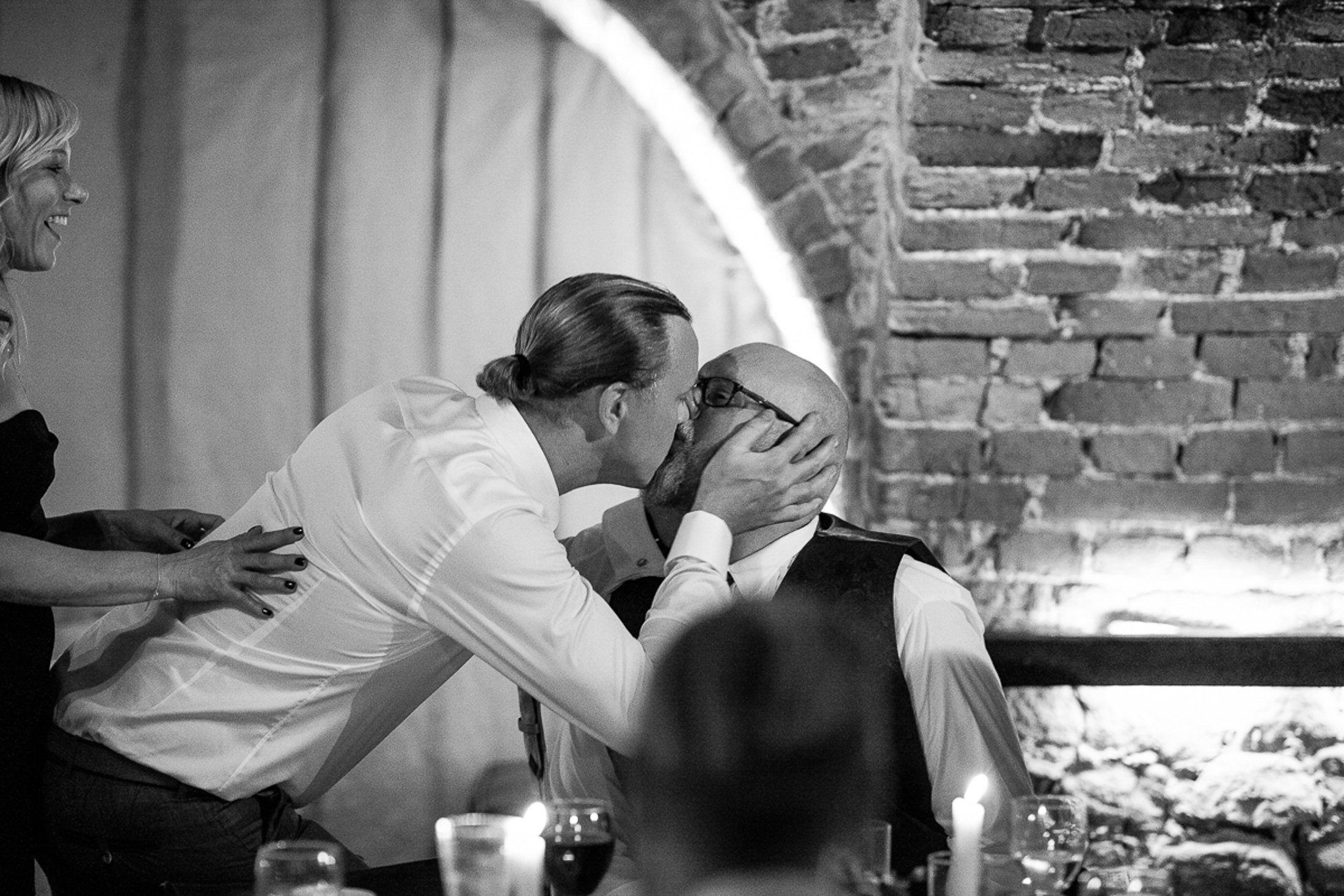 En man klädd i en smal vit skjorta kysser en annan man som sitter ner