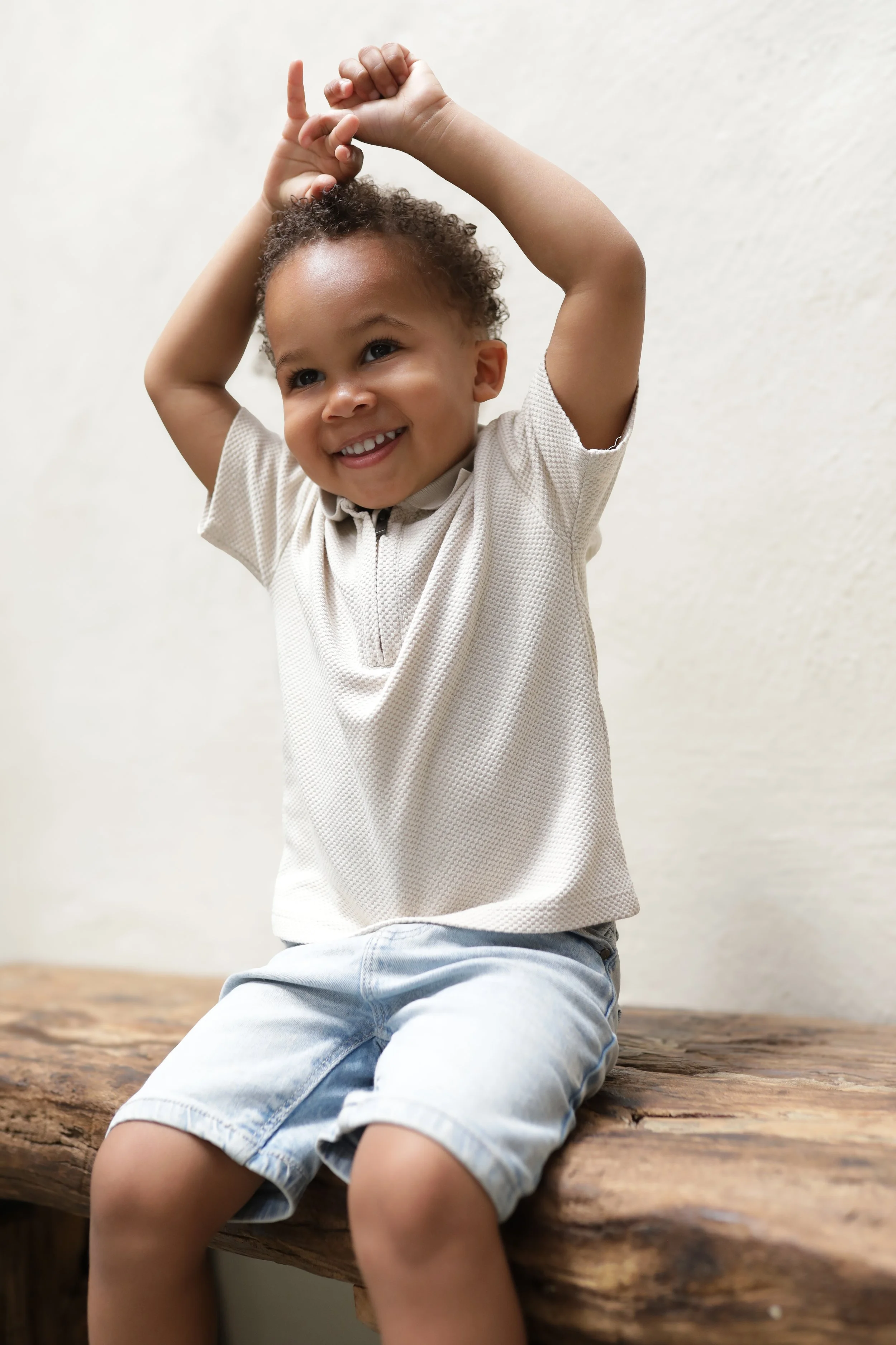 Lachende jonge jongen met krullend haar zittend op een boomstam, gekleed in een beige shirt en korte broeken.