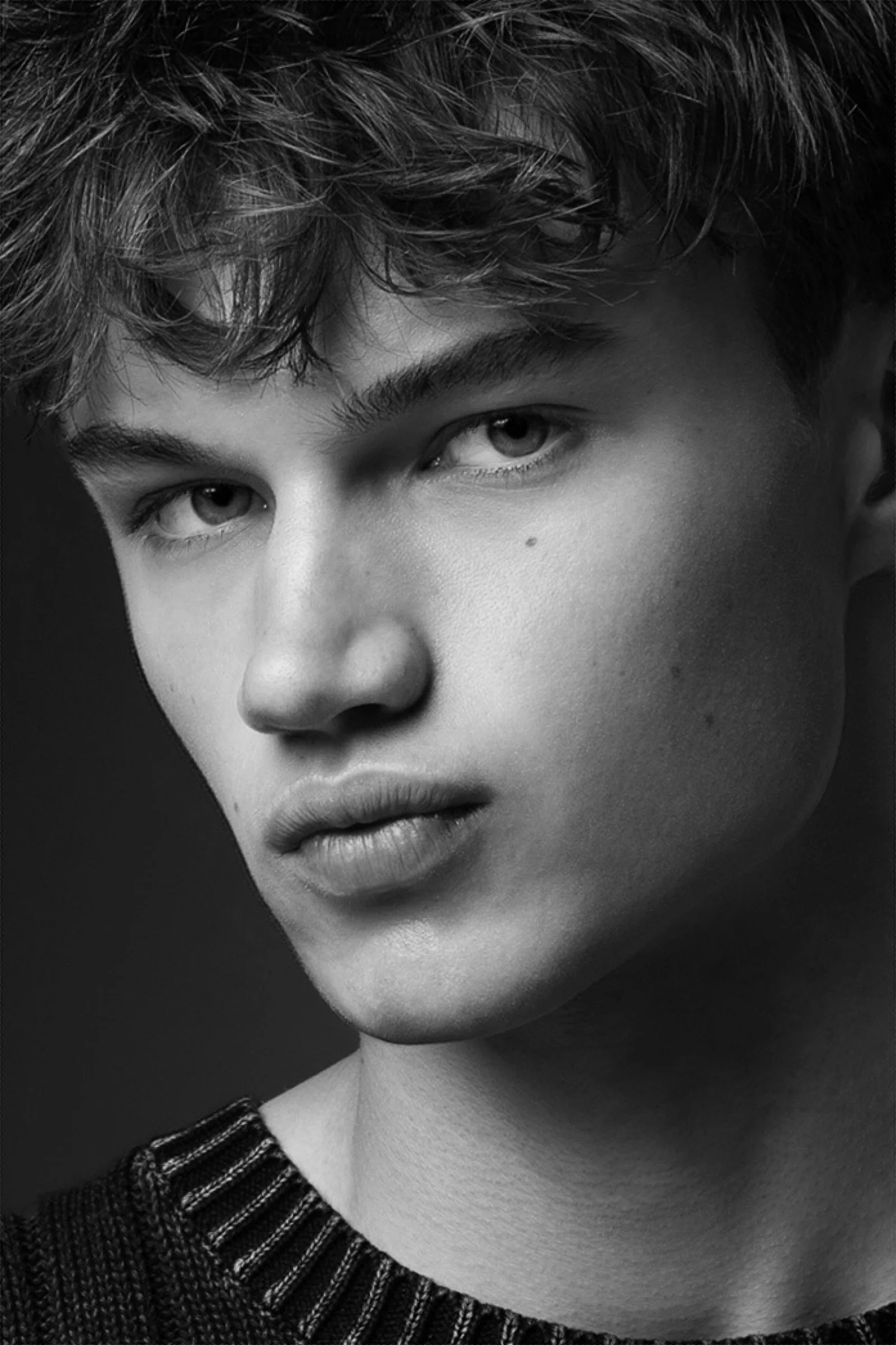 Close-up zwart-wit foto van een jonge man met kort, golvend haar, opvallende blauwe ogen en volle lippen, gekleed in een donker gebreid sweater met strepen.