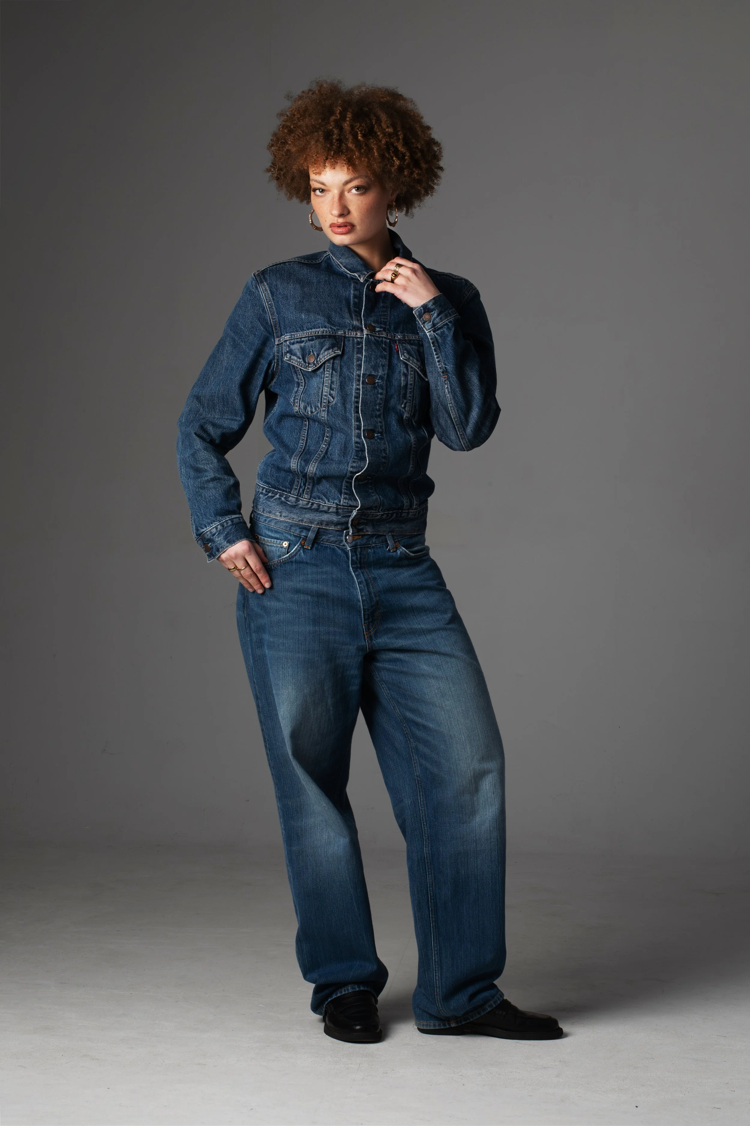 Een vrouw met een krullende haardos draagt een denim jas en broek, poseert tegen een grijze achtergrond.
