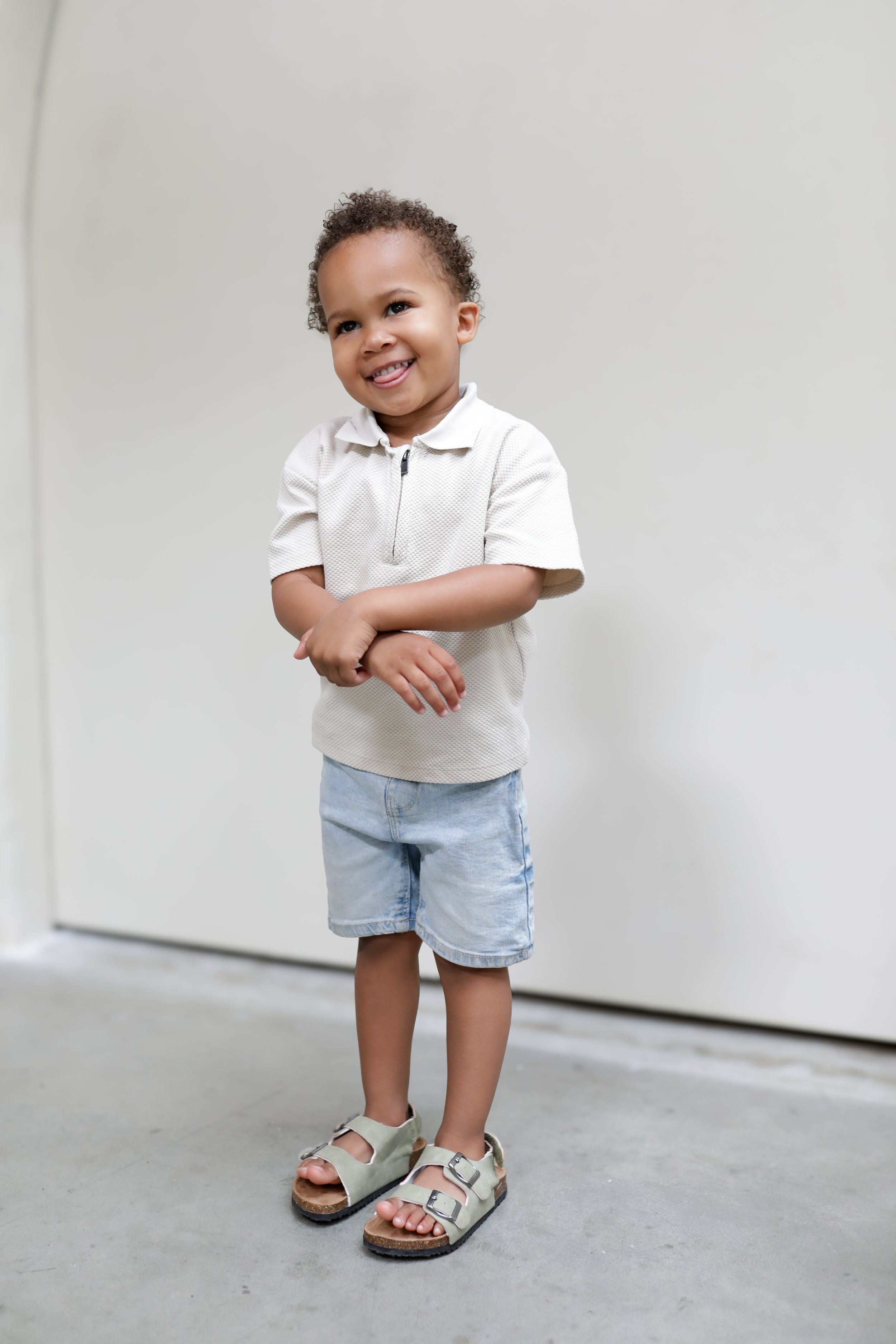 Kleine jongen met gekrulde haar, glimlachend, met armen gekruist, draagt witte polo, korte blauwe shorts en sandalen, staand op een vlakke vloer met een witte muur op de achtergrond.