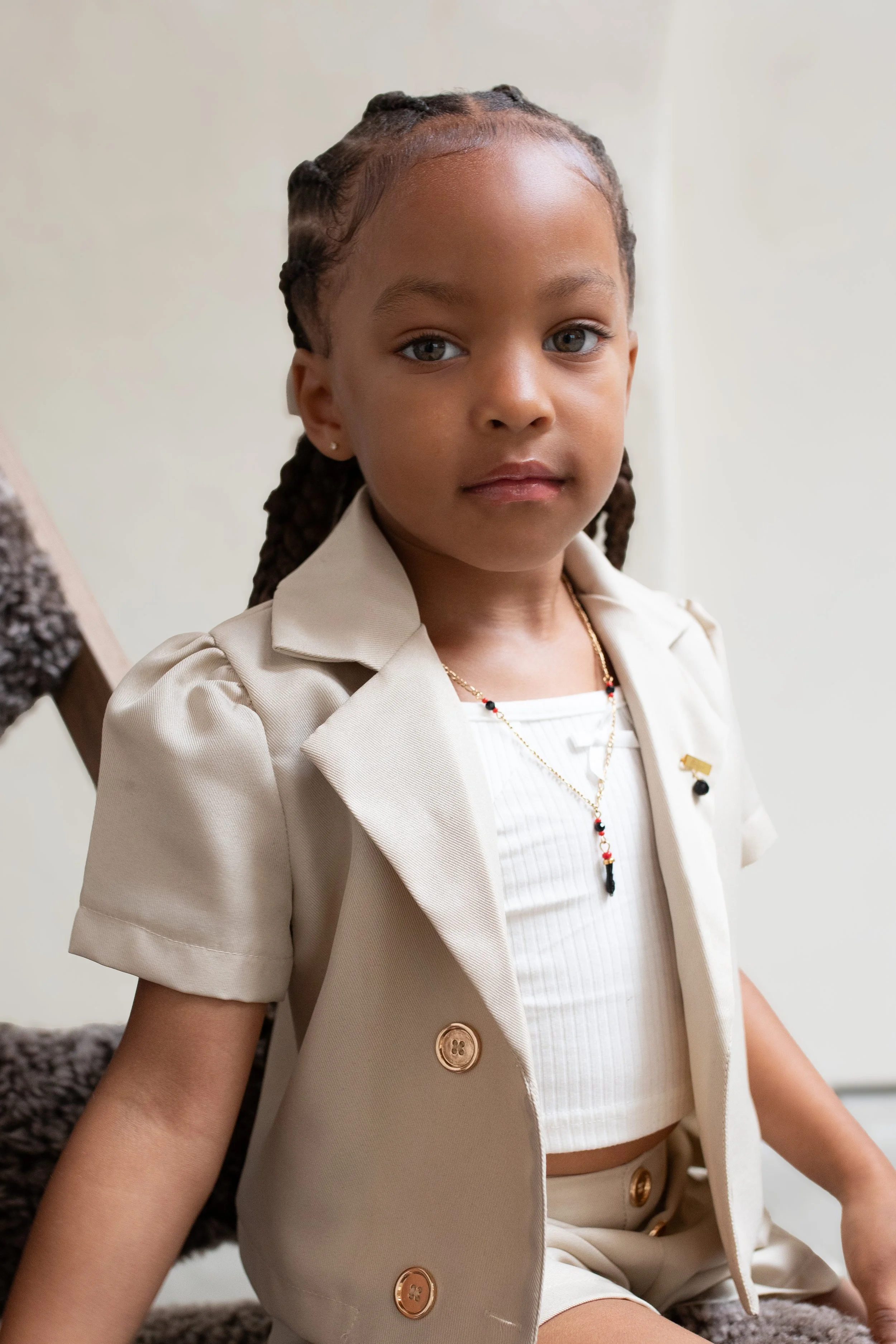 Een jonge meisje met een gevlochten kapsel, zittend op een tapijt, gekleed in een beige blazer en rok, met sieraden om haar nek en oorbellen.