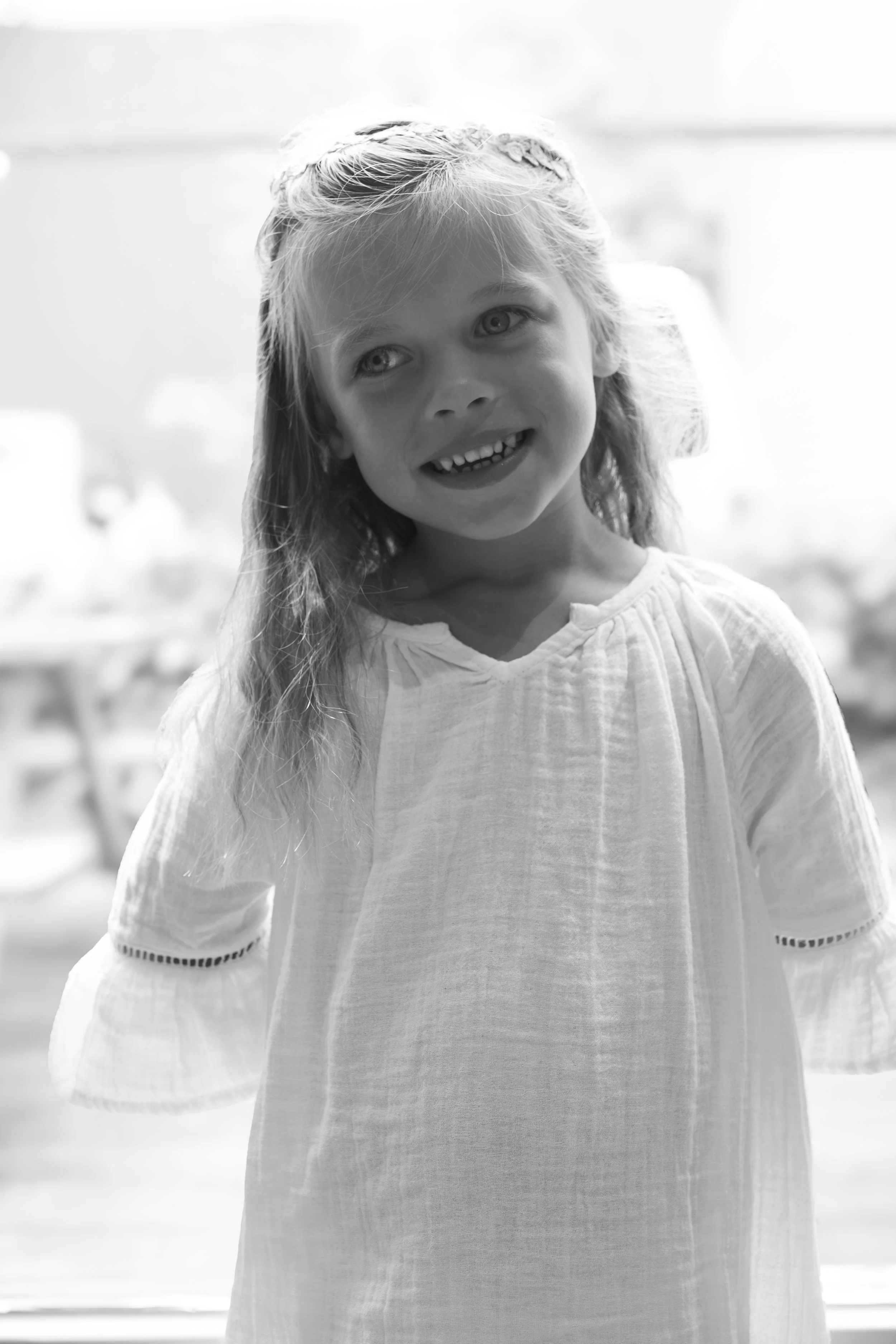 Zwart-wit foto van een glimlachende jong meisje met lang, licht gekruld haar, dat naar rechts kijkt, in een lichte blouse met details aan de mouwen, op een fijne achtergrond. 