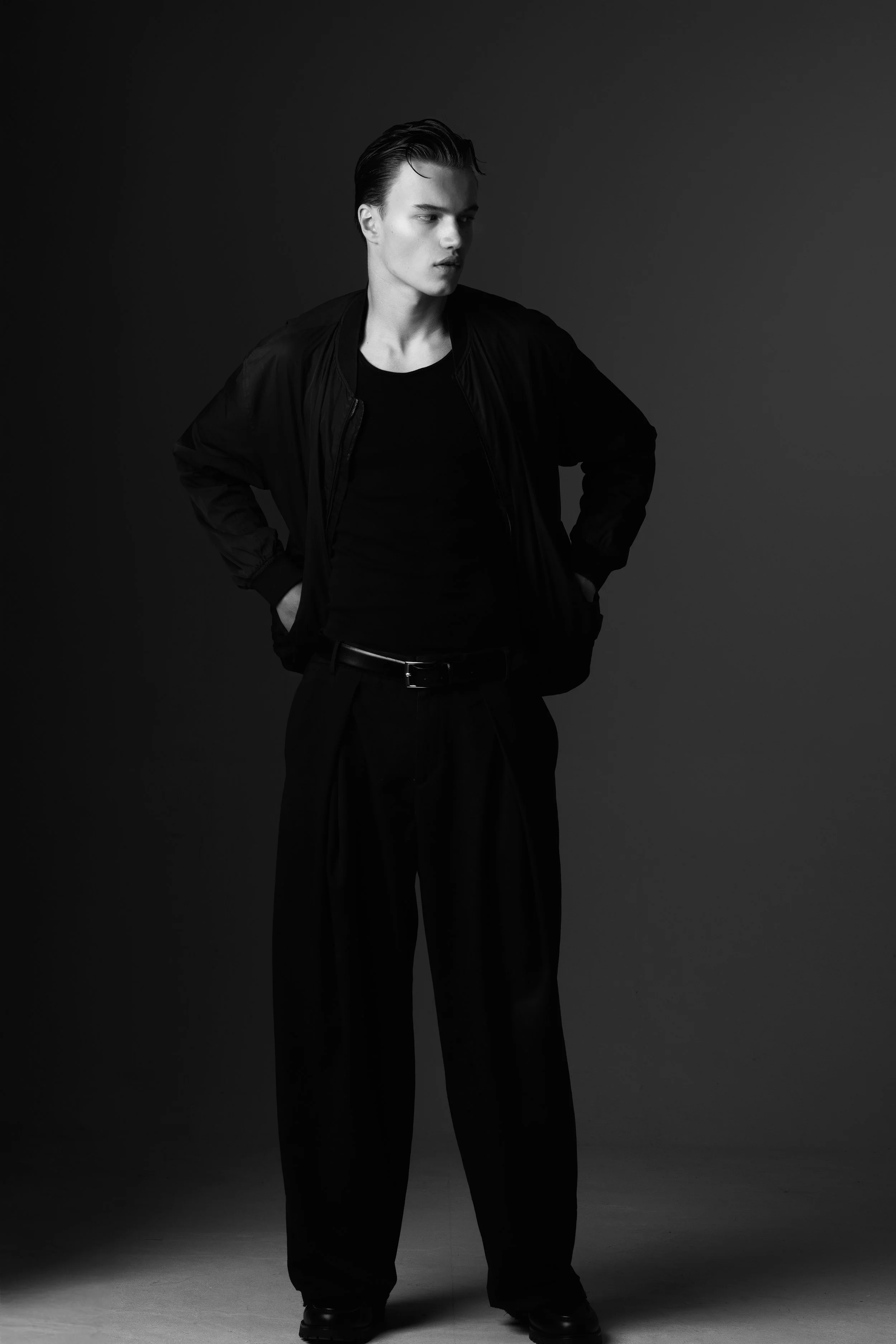 Zwart-wit foto van een jonge man in zwart jack en broek, staand met handen in zij en kijkt naar links, tegen een effen achtergrond.