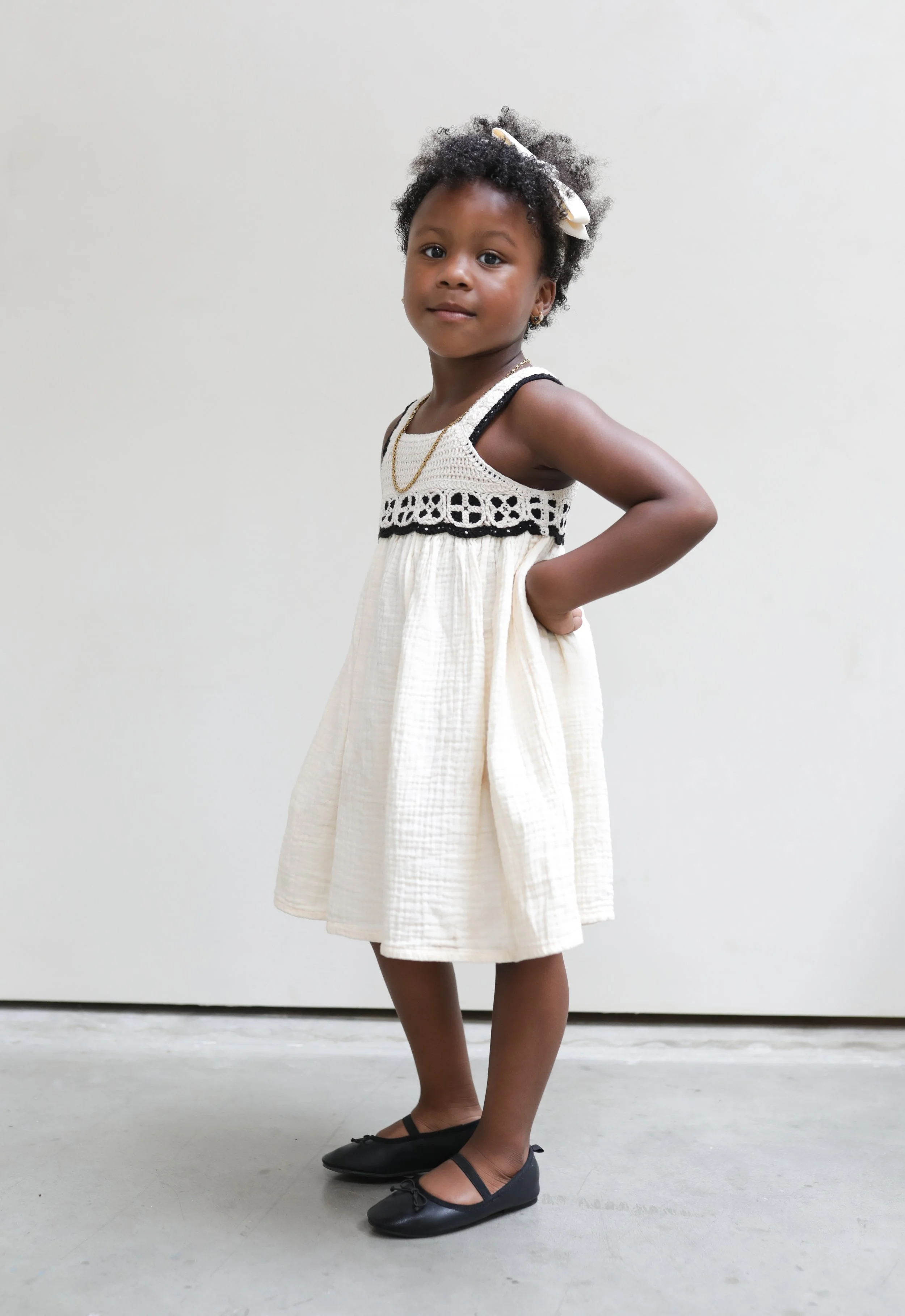Een jonge meisje in een witte jurk met zwarte details, gekleed in zwarte ballerina's, staat met haar rechterhand op haar heup en kijkt naar de camera, tegen een witte achtergrond.