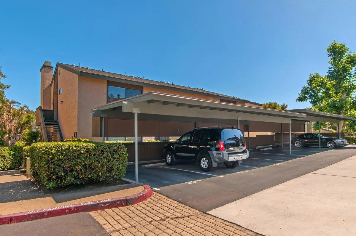 An apartment complex with covered parking spaces, a car parked under the shelter, and a building in the background with stairs leading up to the second floor. The scene is set on a clear, sunny day with blue skies and some green trees.