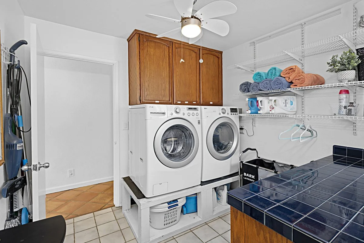 Laundry room with washer and dryer on a raised platform, wooden cabinets above, and wire shelving with folded towels and laundry supplies. A ceiling fan with lights is above, and a tiled countertop is on the right side.