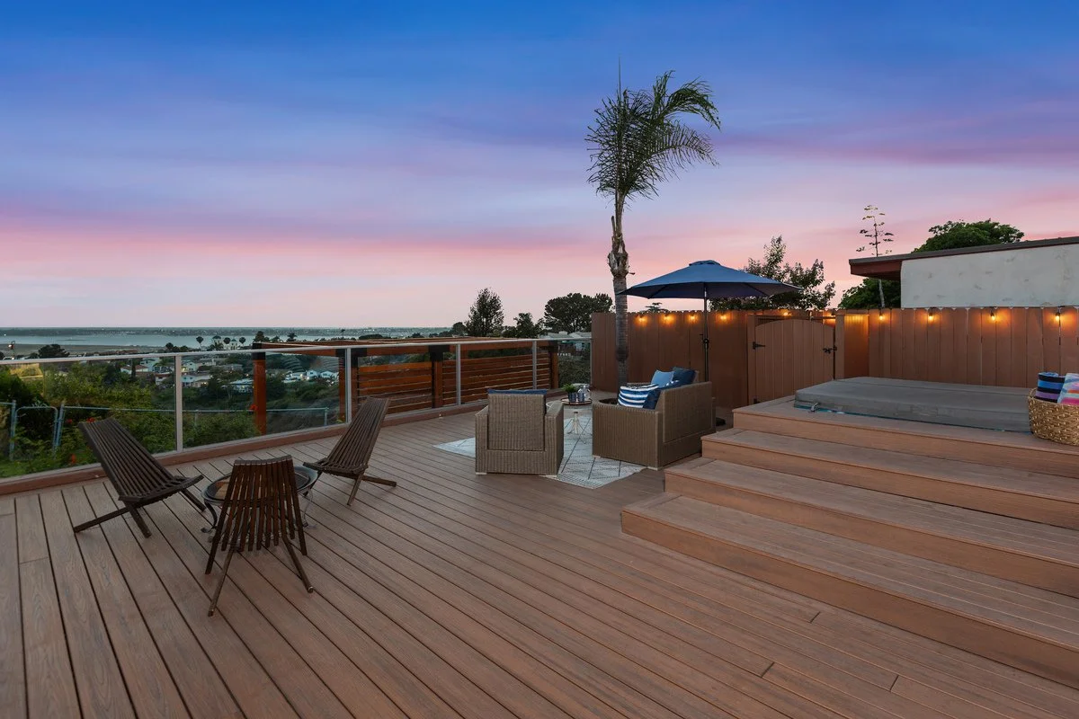 A spacious rooftop deck with outdoor furniture, a hot tub, and a privacy fence, overlooking a scenic view at sunset with colorful sky.