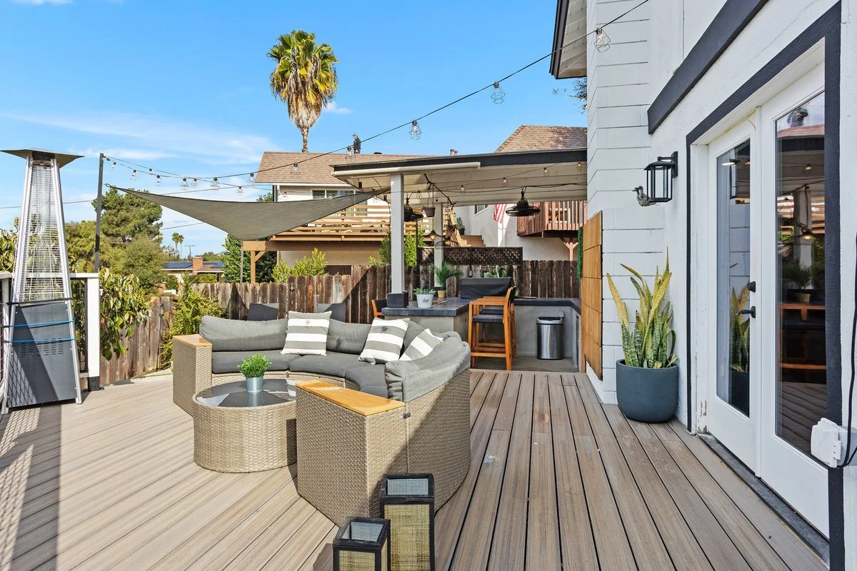 An outdoor deck area with a round wicker sofa, a glass-topped coffee table, and potted plants. There are string lights above and a tall patio heater to the left. The house has white siding, and there are stairs and a barbecue area in the background. 