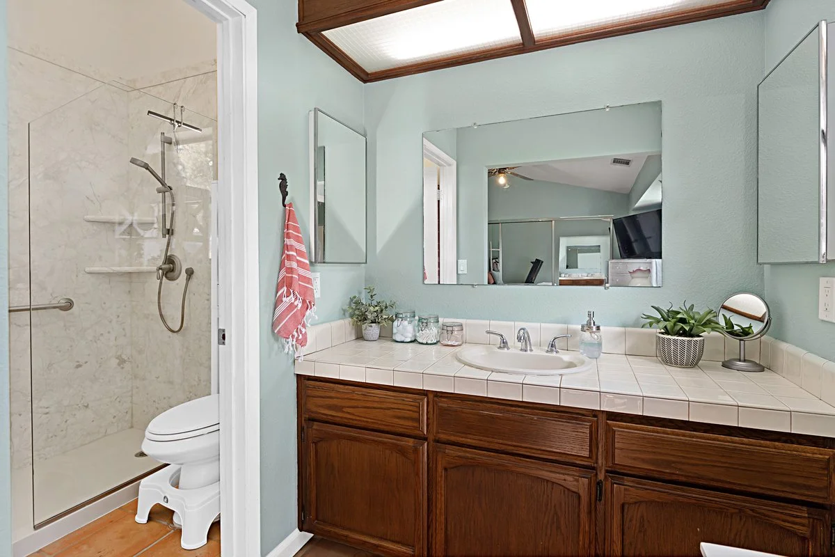 A bathroom with a wooden vanity, a large mirror, and a shower with a glass door. Decor includes small plants and jars on the countertop, and a red and white towel hanging on a hook.