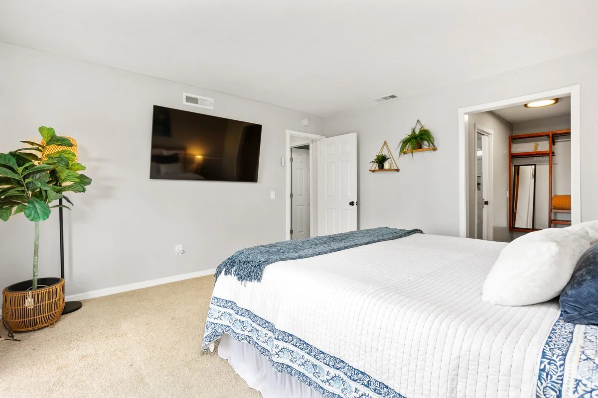 A bedroom with a bed, a wall-mounted TV, a potted plant, and decorative shelves with plants, with an open door leading to a closet.